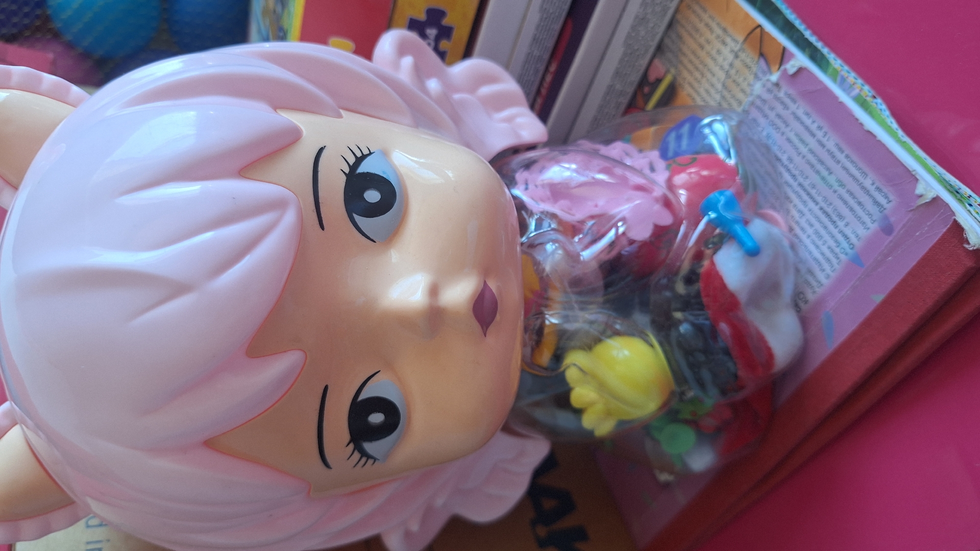 The image shows a close-up of a pink plastic doll head with large eyes and cat ears. The doll is positioned in front of a stack of books. Inside a clear plastic wrapper around the doll's neck and chest, there are small colorful items, including a yellow flower-shaped object and a pink flower. The background is blurred and shows hints of other objects and colors, suggesting an indoor setting. There are no identifiable landmarks or clues that would indicate a specific location in the city of Samara, Russia.