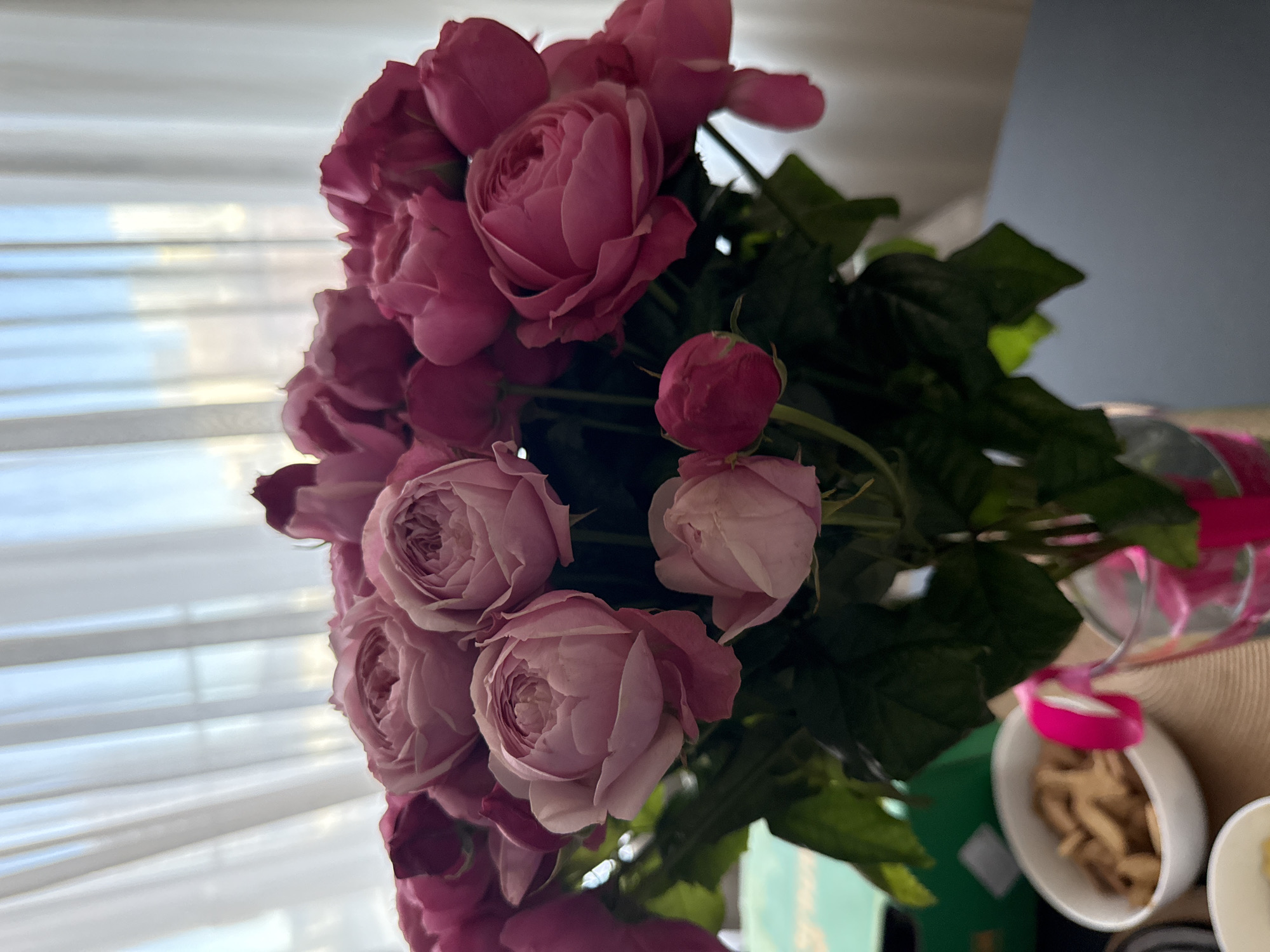 A vertical, close-up photograph captures a vibrant bouquet of pink roses dominating the right and central portions of the frame. The roses display various stages of bloom, from tight buds to fully open blossoms with numerous layered petals, ranging in color from deep fuchsia to softer pastel pink. Lush green foliage is visible among the flowers.
To the left of the bouquet is a bright window, covered by horizontal white blinds that diffuse the natural daylight. A faint light blue hue can be seen through the blinds, suggesting the outdoor sky. In the top right background, a plain light blue-grey wall provides a neutral backdrop.
In the bottom right corner, a section of a surface, likely a table, is visible. On this surface, a white oval bowl holds a collection of light brown, elongated snack items, possibly cookies or crisps. A bright pink decorative element, perhaps a ribbon or a part of the flower's wrapping, is seen near the bowl and extends towards the edge of a clear glass or vase that holds the roses. A textured, light brown woven material, consistent with a placemat or tablecloth, is beneath the bowl. A hint of a green object is also visible in the bottom left of this area.
The scene suggests a serene domestic setting, possibly a relaxed daytime moment in a home in Krasnoyarsk, Russia. The presence of the bouquet and snacks might indicate a celebration, a gift, or simply an aesthetic arrangement. The natural light creates a soft and inviting atmosphere. No people or visible text are present in the image.