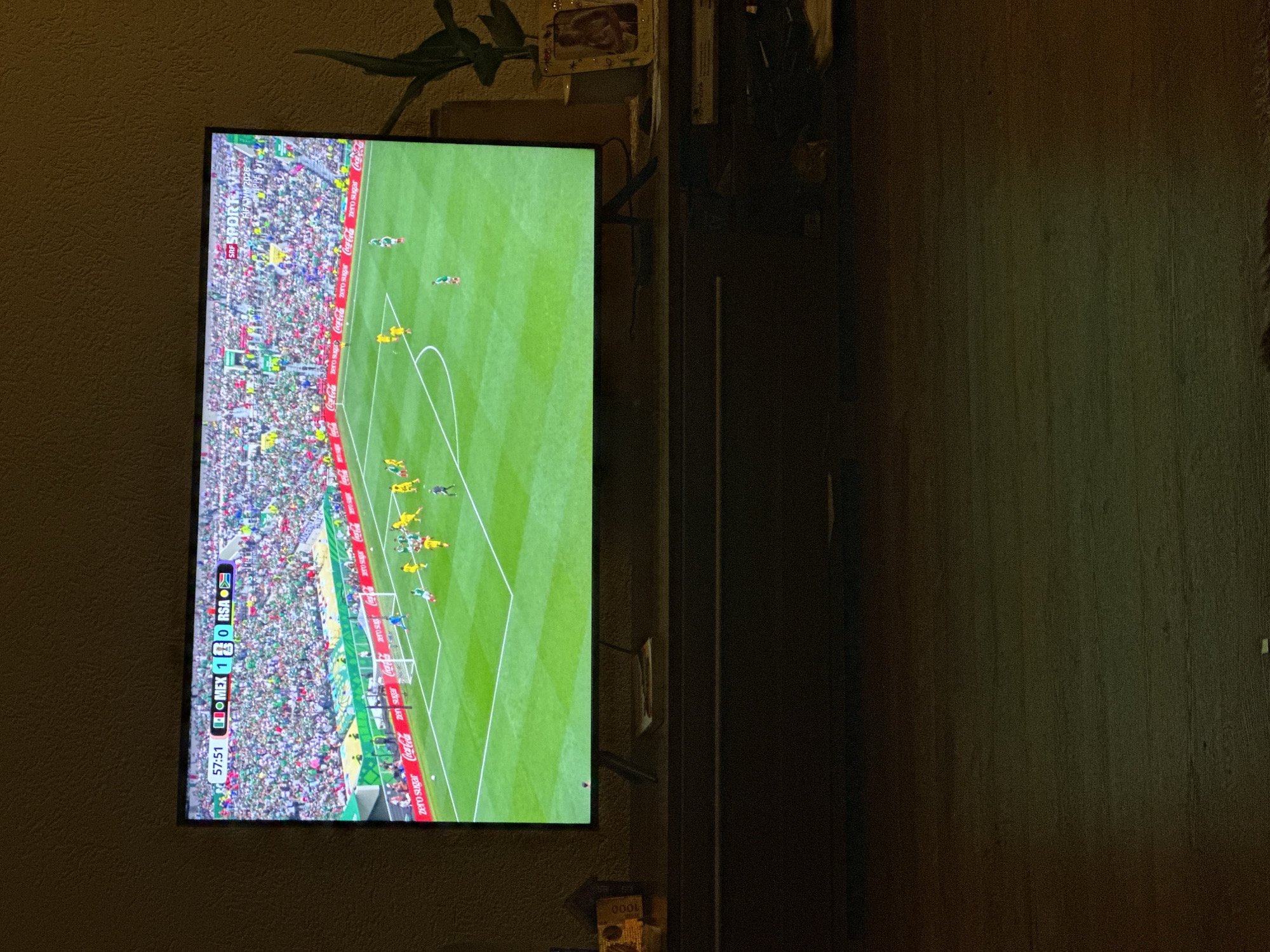 The image shows a television screen displaying a soccer match. The score shown on the screen is 57:51, with Mexico leading 1-0 against RSA. The stadium is filled with spectators, and there are various advertisements around the field, including Coca-Cola. The television is placed on a dark wooden cabinet. To the right of the television, there is a potted plant and a framed picture. The overall environment appears to be a living room or entertainment area. There is no information in the image to indicate the location in the city of Hüsen, Switzerland.