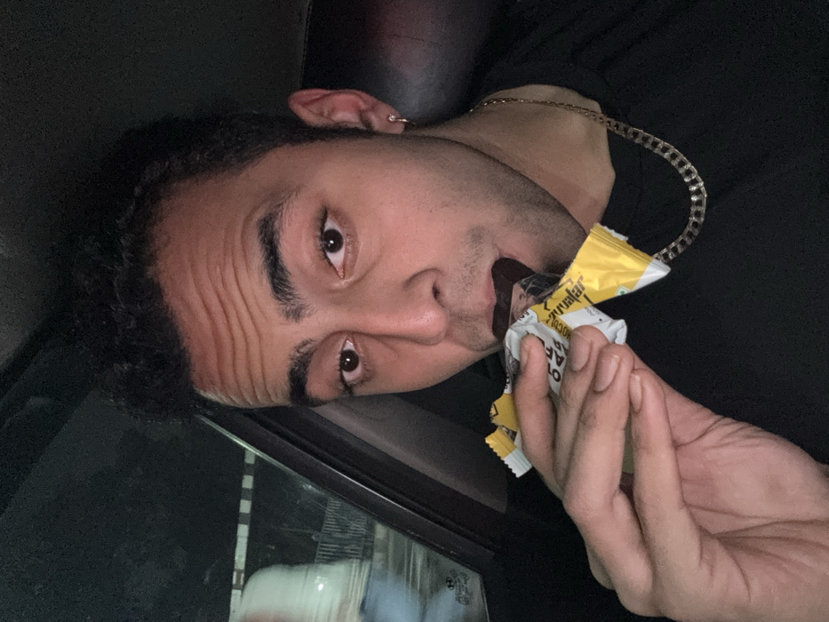 A male individual with dark hair and brown eyes is depicted in a close-up, chest-up shot inside a vehicle. He wears a dark, short-sleeved top and a gold-colored chain necklace; a small silver-toned hoop earring is present in his left ear. Prominent horizontal wrinkles are visible on his forehead. His right hand holds a partially unwrapped food bar, which he is actively biting into. The bar's wrapper is white and yellow, with legible text fragments including "AVATAR," "Chocolate," "OATS," and "45G." Through the vehicle's window on the left, a blurred, illuminated white dome structure with horizontal architectural features below is visible, positively identifying the location as the vicinity of the Victoria Memorial in Kolkata, India. The ambient lighting is dim, indicating a nighttime setting.