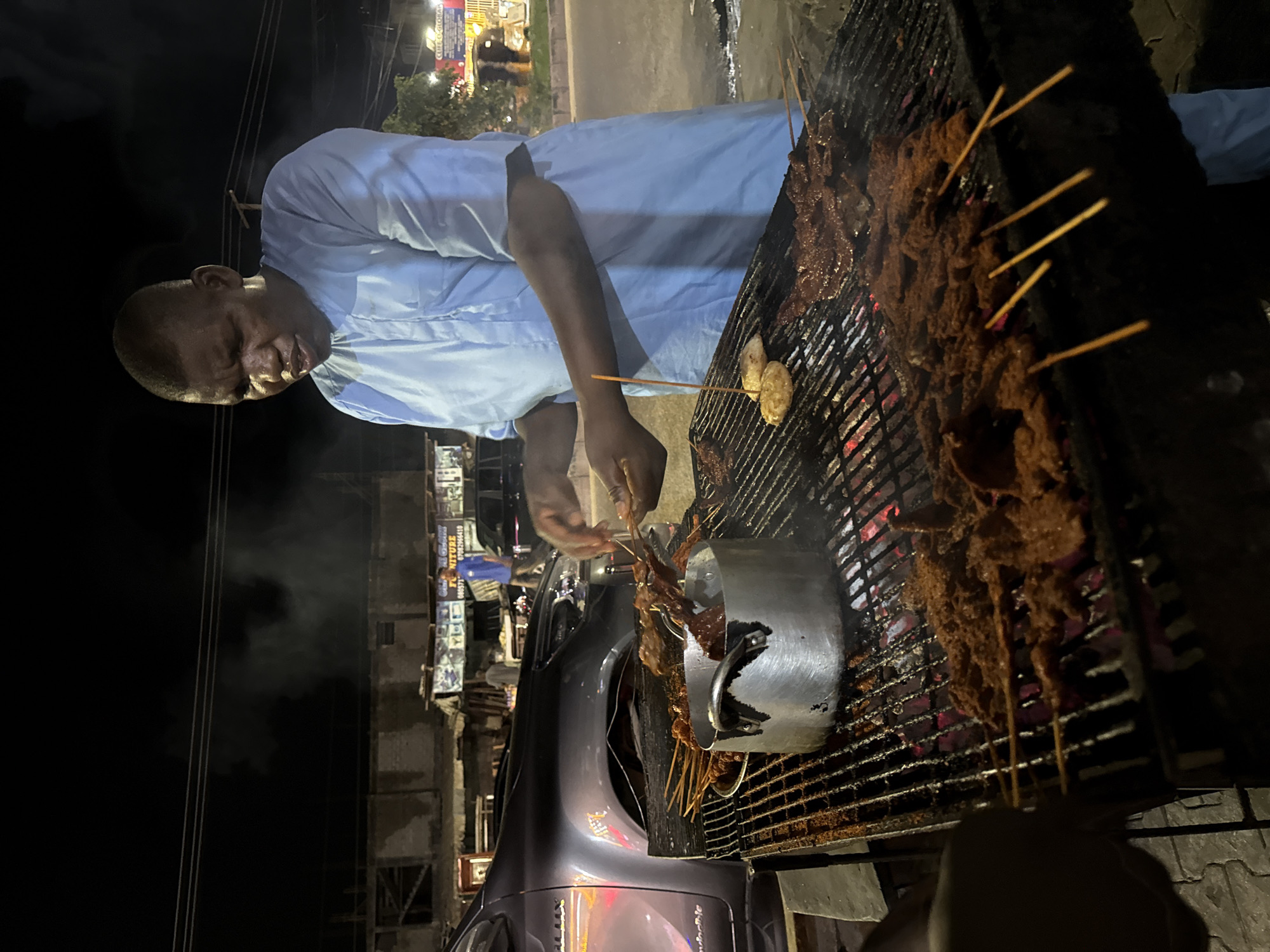 In Ado, Nigeria, a man dressed in a light-colored, long-sleeved shirt stands next to a grill at night. The grill is loaded with skewered meat; some are cooking on the grill, and others are being placed into a metal pot next to it. A car is visible in the foreground. Streetlights and buildings line the background.