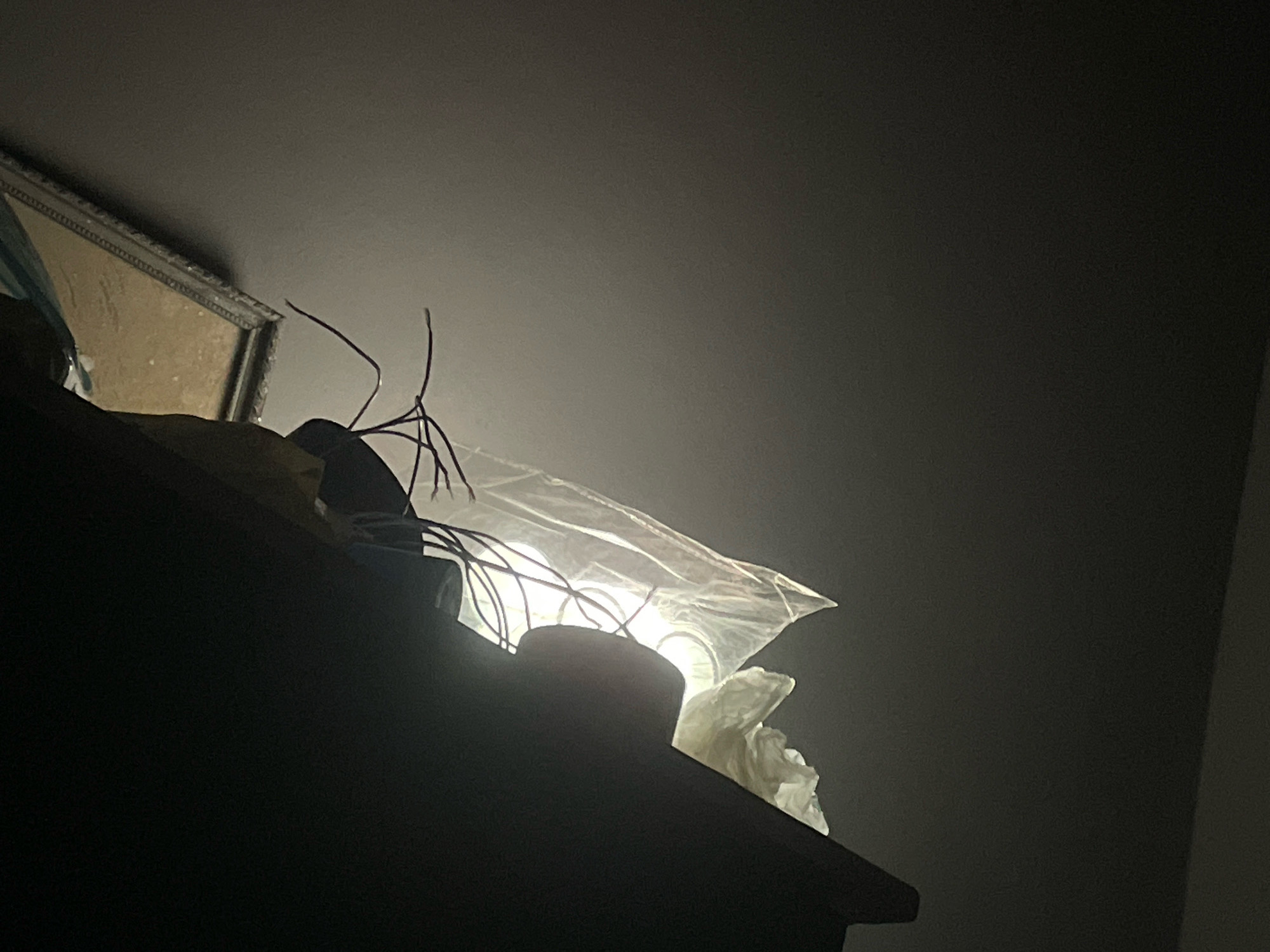 The image shows a dimly lit interior scene, likely a room in Nukus, Uzbekistan. In the foreground, the top of a dark, wooden piece of furniture is visible, suggesting a shelf or cabinet. Resting on this surface are several objects. A bright light source, possibly a lamp with a translucent or white material covering it, is prominently displayed. The light illuminates thin, dark, wire-like structures that appear to be entangled or arranged around the light source. To the left, a portion of a framed picture or mirror with an ornate, textured frame is visible, angled upwards. The background consists of a plain, dark wall or ceiling. The overall impression is one of a cluttered, somewhat artistic or makeshift arrangement in a low-light setting. No people are present in the image.
