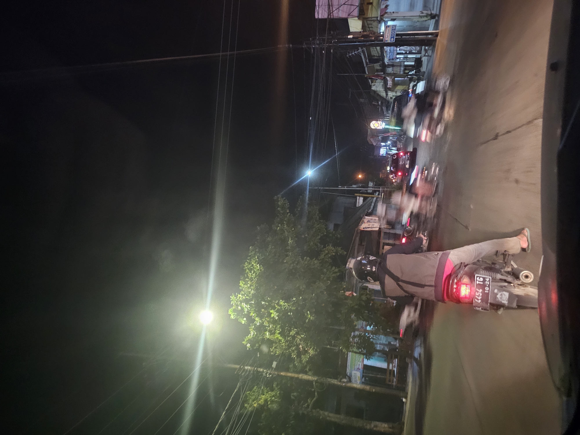The image depicts a street scene at night in Lamongan, Indonesia. A person is riding a motorcycle in the foreground, facing away from the camera. The motorcycle has a license plate that reads "B 4454 TG 10-24". The rider is wearing a helmet and a jacket. Other vehicles, including cars and motorcycles, are visible on the road, some of which are blurred due to motion. Streetlights illuminate the scene, casting light on the road and surrounding buildings. Trees and power lines are also present. Shops and storefronts line the side of the street, with some signs and advertisements visible. The overall impression is a busy urban environment at night.