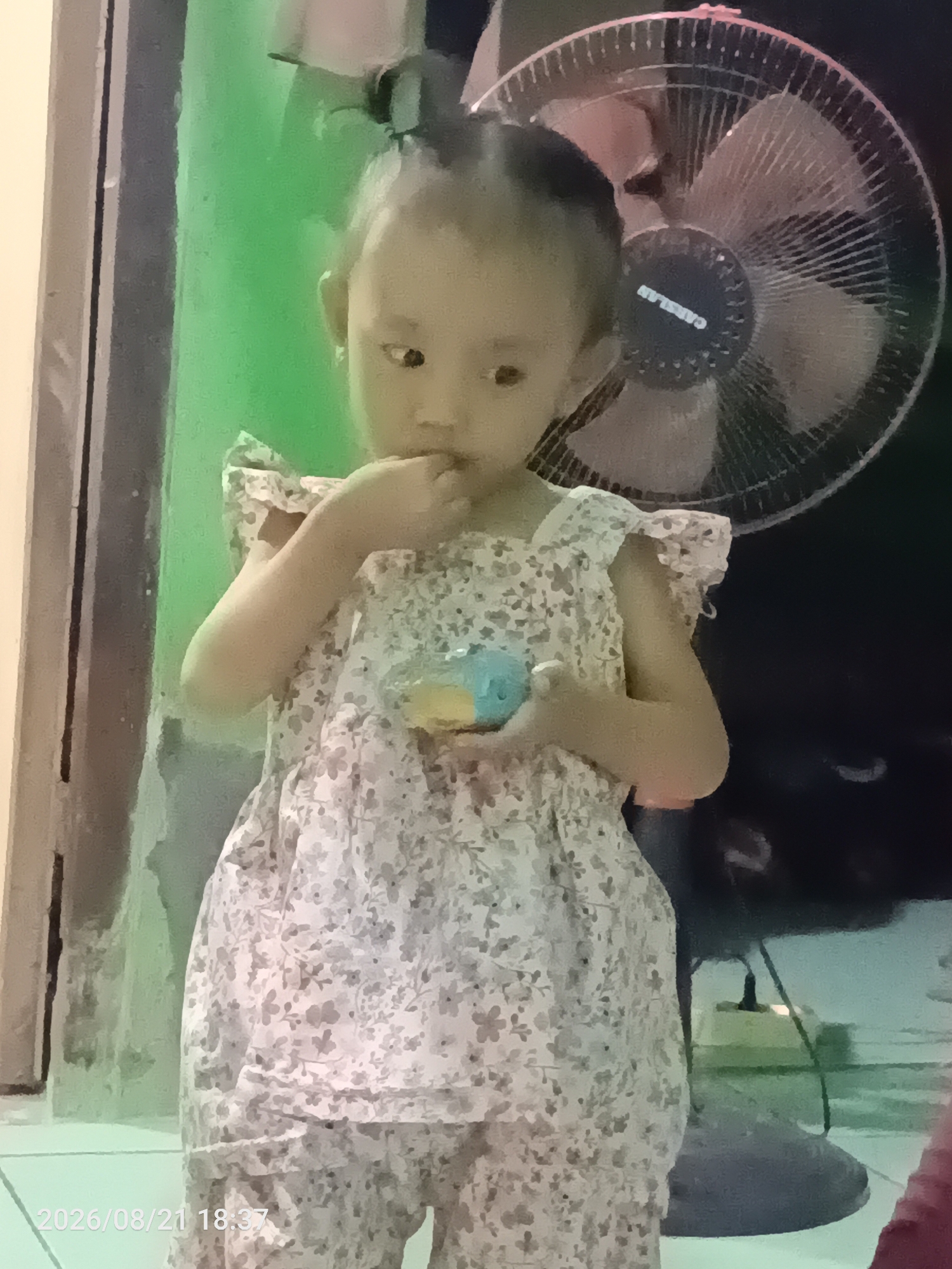 A young child wearing a floral patterned dress and pants stands in front of a fan. The child is holding a small blue and yellow object in their right hand and has their left hand near their mouth. The background is dark with some green lighting on the left side. A date stamp "2026/08/21 18:37" is visible in the bottom left corner. The setting appears to be indoors, possibly a home in the city of Jawilan, Indonesia.
