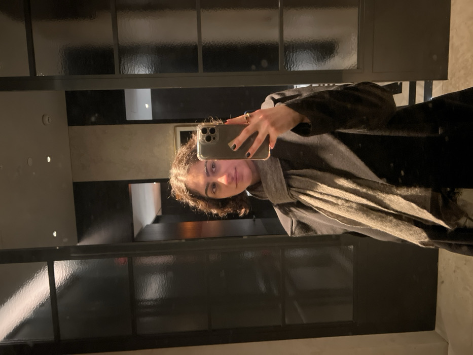 A woman takes a mirror selfie, holding her phone up. She has curly brown hair, light skin, and wears a dark coat with a grey scarf. Her nails are painted black. The background features black-framed mirrored panels above and a dark, narrow hallway on the side. The lighting is dim, creating shadows.