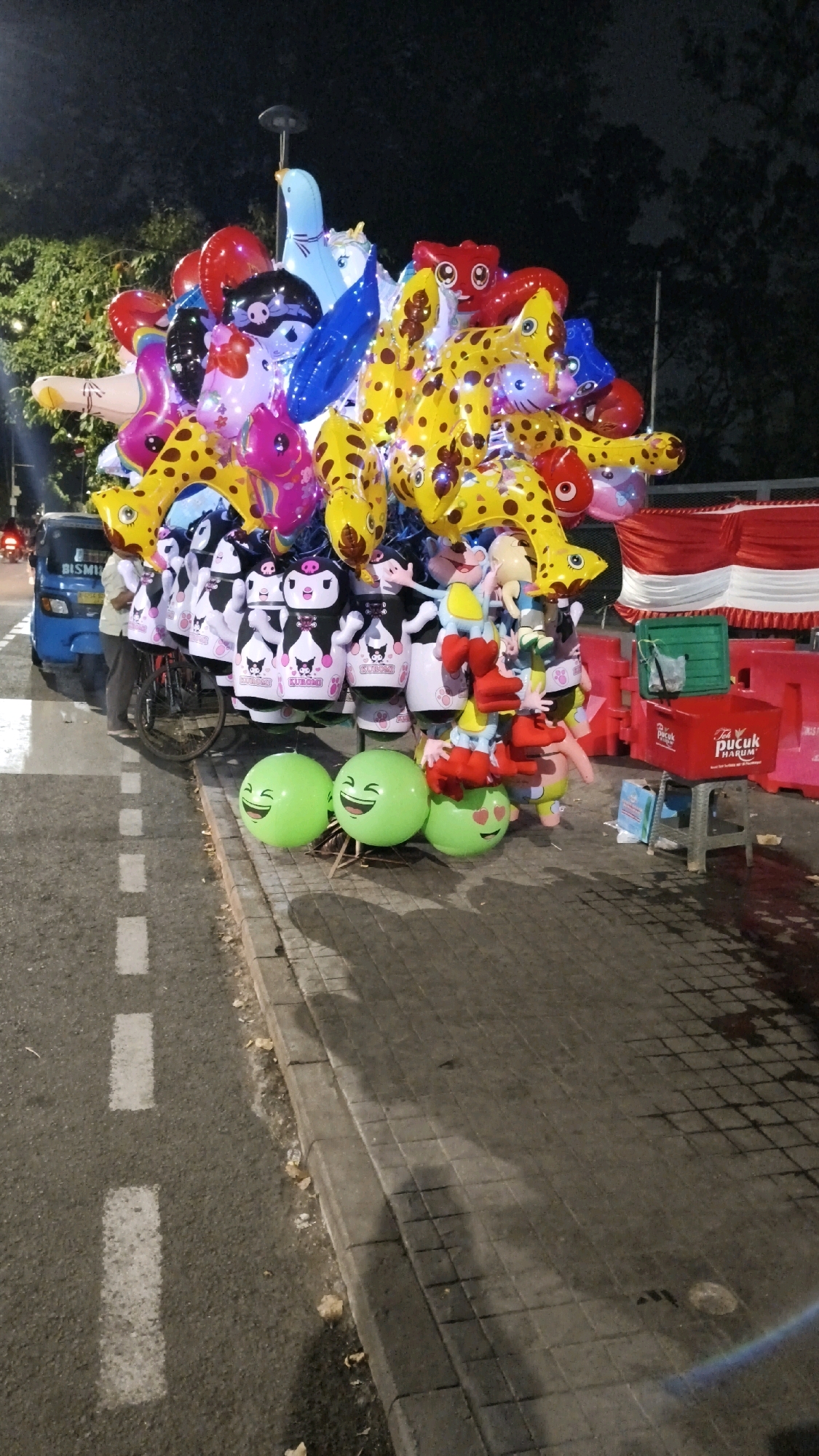 The image depicts a nighttime street scene in South Jakarta, Indonesia, with a vendor selling balloons. A large cluster of colorful balloons, including animal shapes and cartoon characters like Kuromi, is prominently displayed. Some balloons are in the shape of smiling green faces. The balloons are attached to a bicycle, suggesting the vendor is mobile. A blue motorized tricycle is parked nearby. In the background, red and white striped fabric is visible, possibly part of a stall or decoration. The ground is paved with asphalt and cobblestones, with white road markings. The scene is dimly lit, with streetlights and ambient city lights illuminating the area. The overall impression is a lively street-side market atmosphere.
