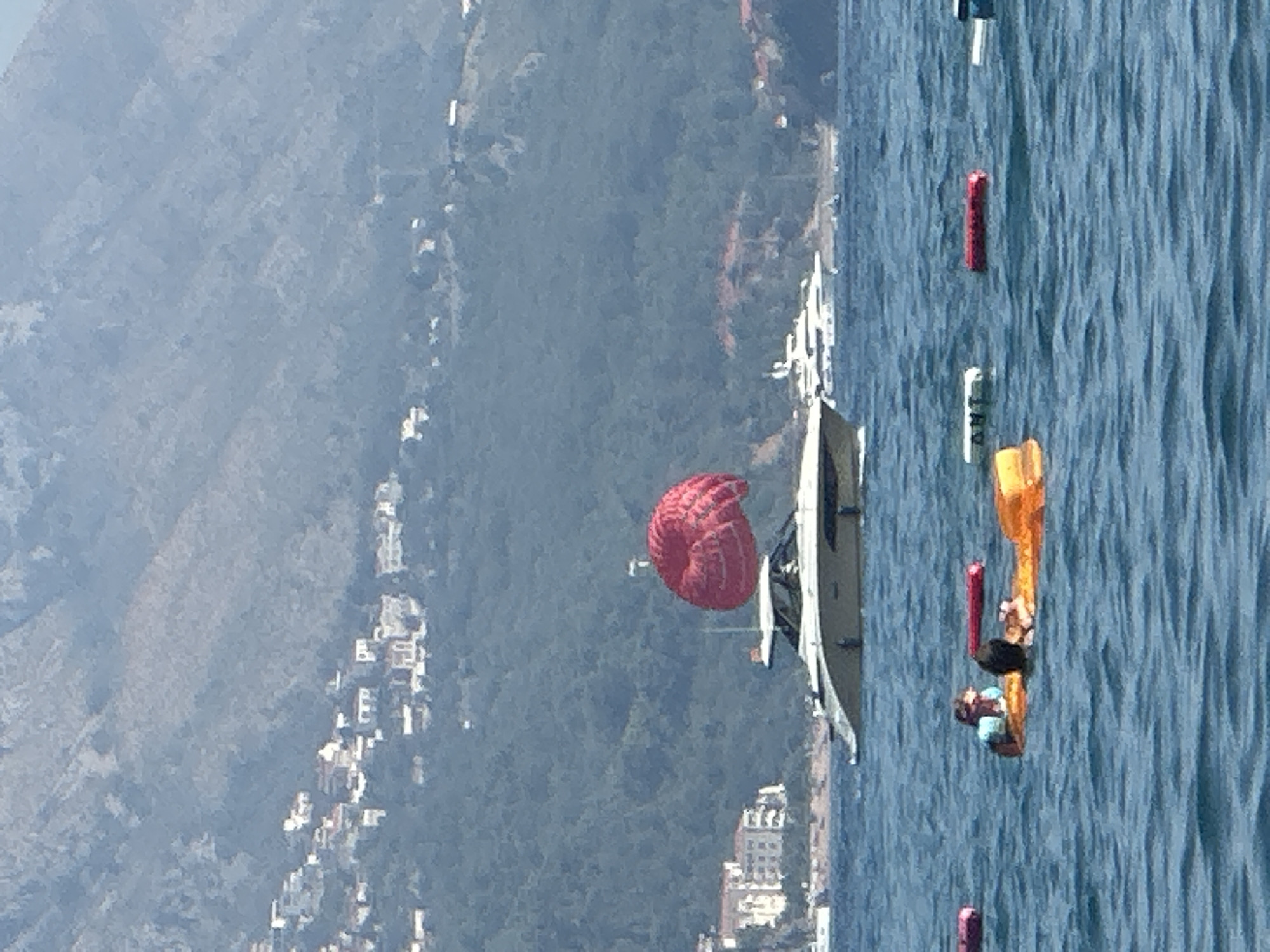 In the city of Boreti, Montenegro, a parasail with a vibrant red canopy is suspended in the air above a white and gold yacht navigating the blue water. The yacht is positioned in the middle ground, with a hillside dotted with buildings and trees rising behind it. In the foreground, two individuals are partially submerged in the water, resting on an orange inflatable raft. Several buoys, some red and some white, are scattered across the water's surface. The scene is set against a backdrop of a hazy, mountainous landscape.