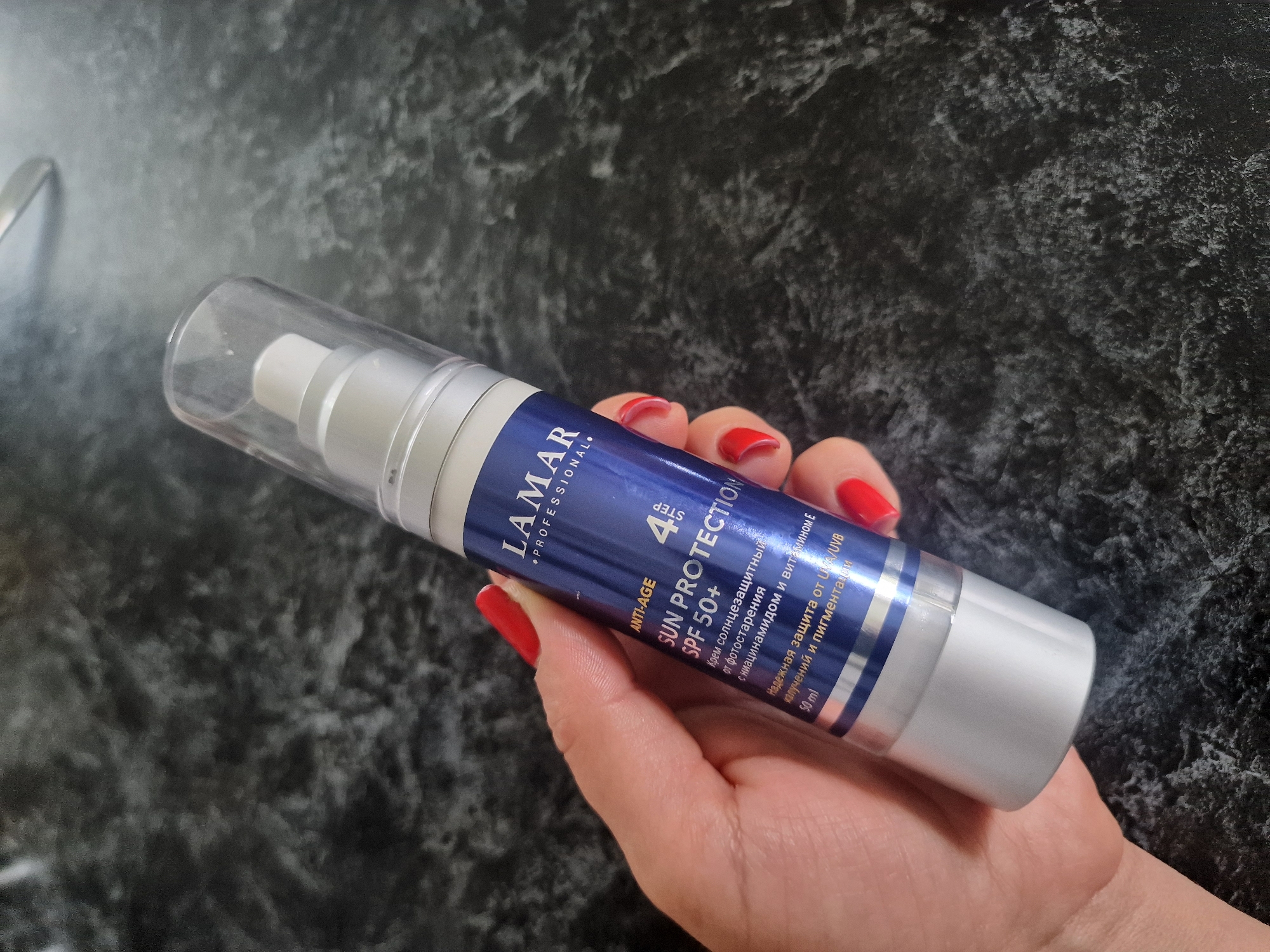 In this close-up, medium shot, a person's hand with bright red nail polish holds a bottle of "LAMAR PROFESSIONAL. ANTI-AGE SUN PROTECTION SPF 50+". The bottle is silver and blue, with text in English and what appears to be Russian. The text indicates the product is a sunscreen cream offering reliable UVA/UVB protection from photoaging, and it contains niacinamide and vitamin E. The volume is listed as 50 ml. The background is a dark, mottled surface, possibly a countertop. The lighting suggests an indoor setting, potentially during the day given the natural skin tones. The hand's grip and the presentation of the product imply a casual demonstration or product placement. There are no strong indicators of the specific location within Kursavka, Russia, beyond the provided context.