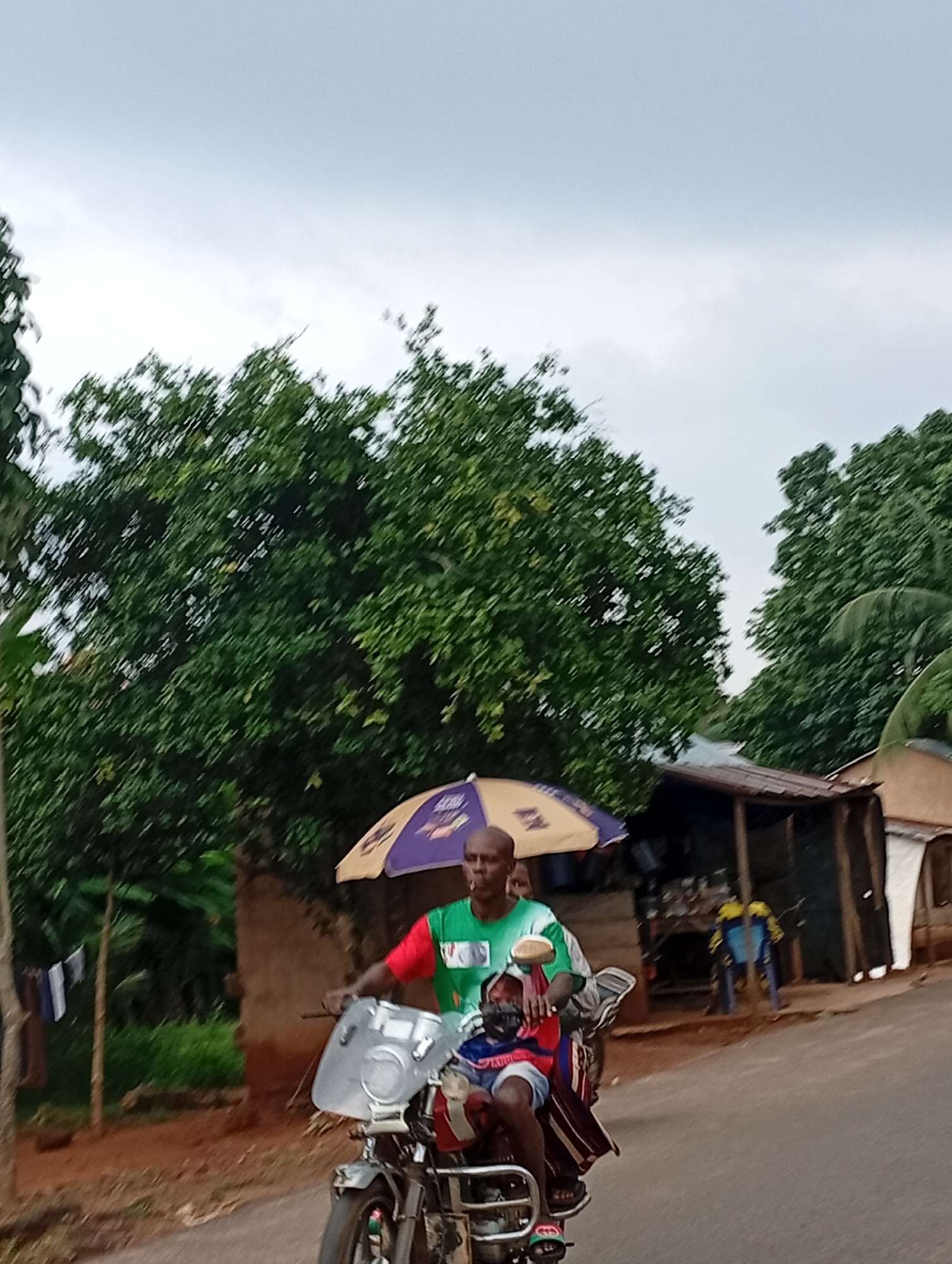 The image shows a man and a child riding a motorcycle in Nsugbe, Nigeria. The man is in the driver's seat, and the child is seated in front of him. Both are wearing casual clothing. The man has an umbrella over them for shade. The motorcycle has a windshield. In the background, there are several small structures, likely shops or homes, with corrugated metal roofs. Green trees and foliage are visible around the buildings and along the side of the road. The sky is overcast. The road appears to be paved.