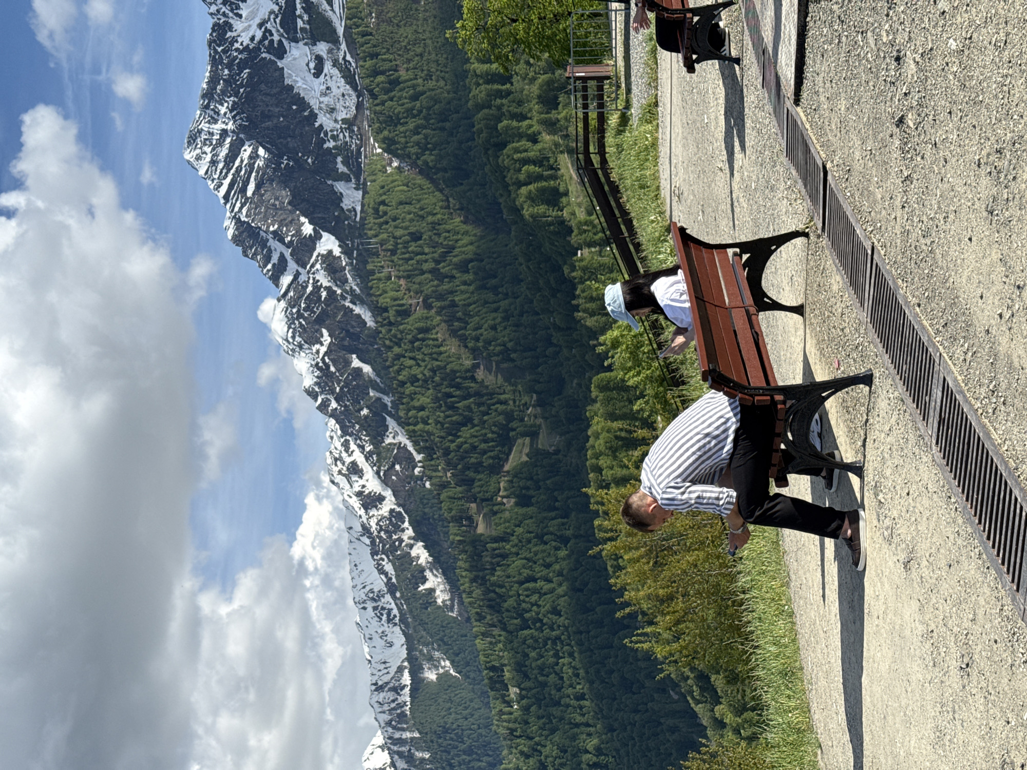 The image displays a scenic mountain landscape observed from a paved pathway, likely in Estosadok, Russia. In the foreground, a light-colored paved area, possibly a gravel path, features a long metal drain grate running parallel along its right side. Several dark-framed wooden benches are placed along this path.
In the midground, beyond a low railing, a vast expanse of dense green coniferous forest covers undulating hills and valleys, receding towards the majestic mountains. The background is dominated by rugged, snow-capped mountains under a bright blue sky with scattered white clouds. Patches of white snow are visible on the dark, rocky peaks and slopes.
Three individuals are present on the path. On the left, a man wearing a striped shirt and dark trousers is bending over, seemingly looking at a phone or something on the ground next to a bench. Further along the path, another person, wearing a light top and a hat, is seated on a bench, facing away from the camera and towards the mountain view. Part of a third person is visible near another bench in the upper right.
The scene is illuminated by bright, direct sunlight, indicating a clear, sunny daytime. No visible text is discernible in the image.