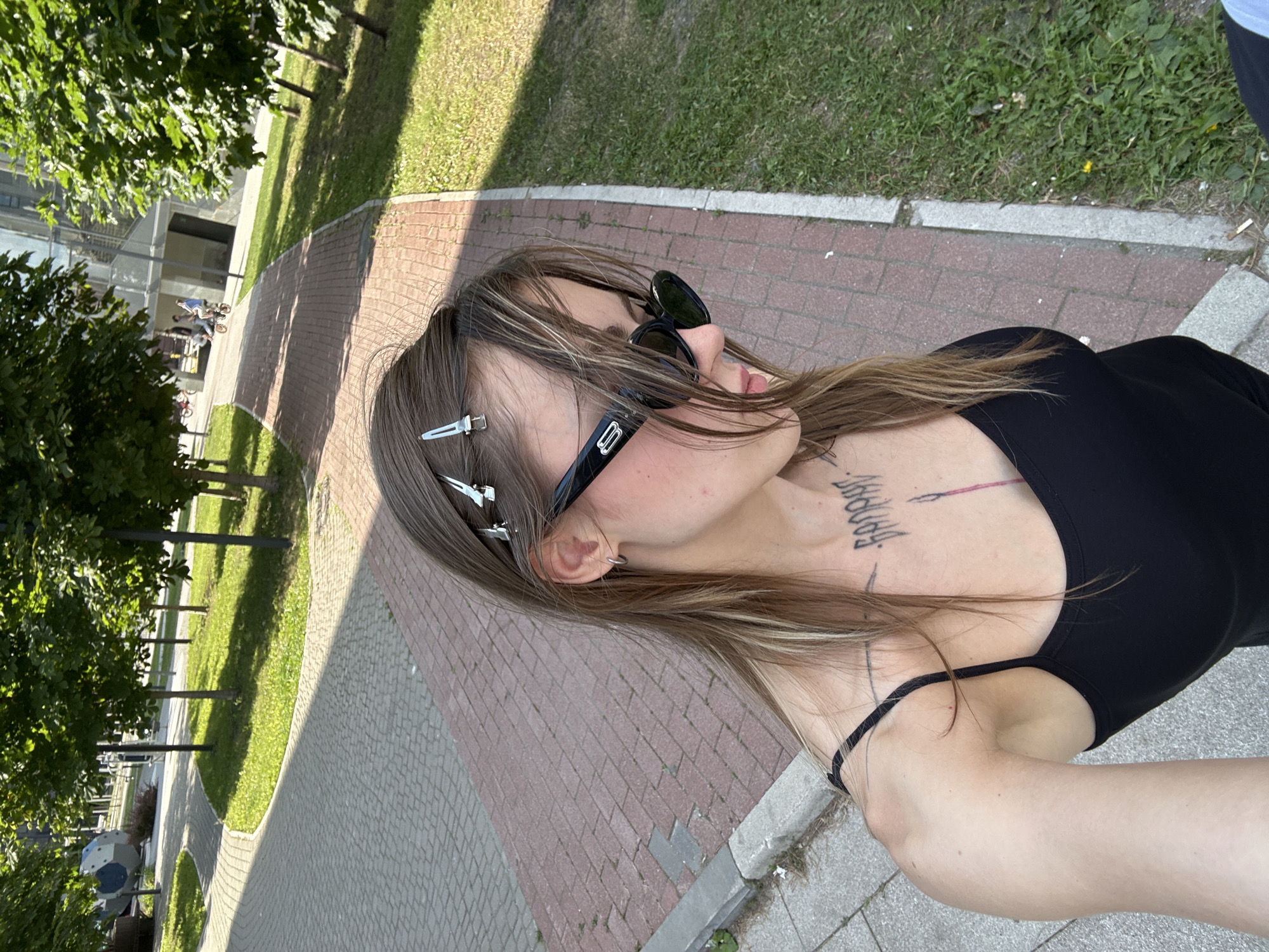 The image shows a close-up, low-angle shot of a young woman from the chest up, posed outdoors. She is wearing a black tank top and black sunglasses. Her hair is long, brown with blonde highlights, and held back with silver barrettes. She has a tattoo on her chest, which appears to be text. The background suggests an urban park setting with trees, grass, and paved pathways made of reddish-brown brick. Sunlight casts shadows, indicating it's daytime. The woman's head is turned away from the camera, and her expression is neutral. The overall atmosphere is casual and sunny. Some text is visible on the woman's tattoo, but it is difficult to decipher clearly. The setting, with its trees and park-like pathways, could be interpreted as a public space in Moscow, Russia, as per the location context provided. There are no other people clearly visible in the immediate foreground, though some blurred figures on bicycles can be seen in the distance on a pathway.