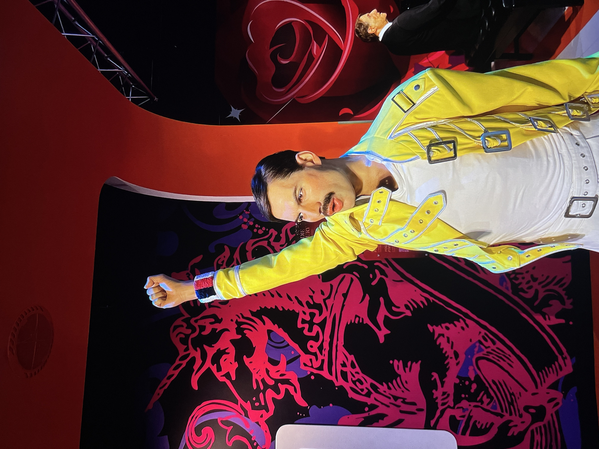 A wax figure of Freddie Mercury in a yellow jacket and white shirt is standing in a display. The wax figure is in front of a red and black patterned wall. Another wax figure is behind Freddie Mercury. The location is in an unidentified city.