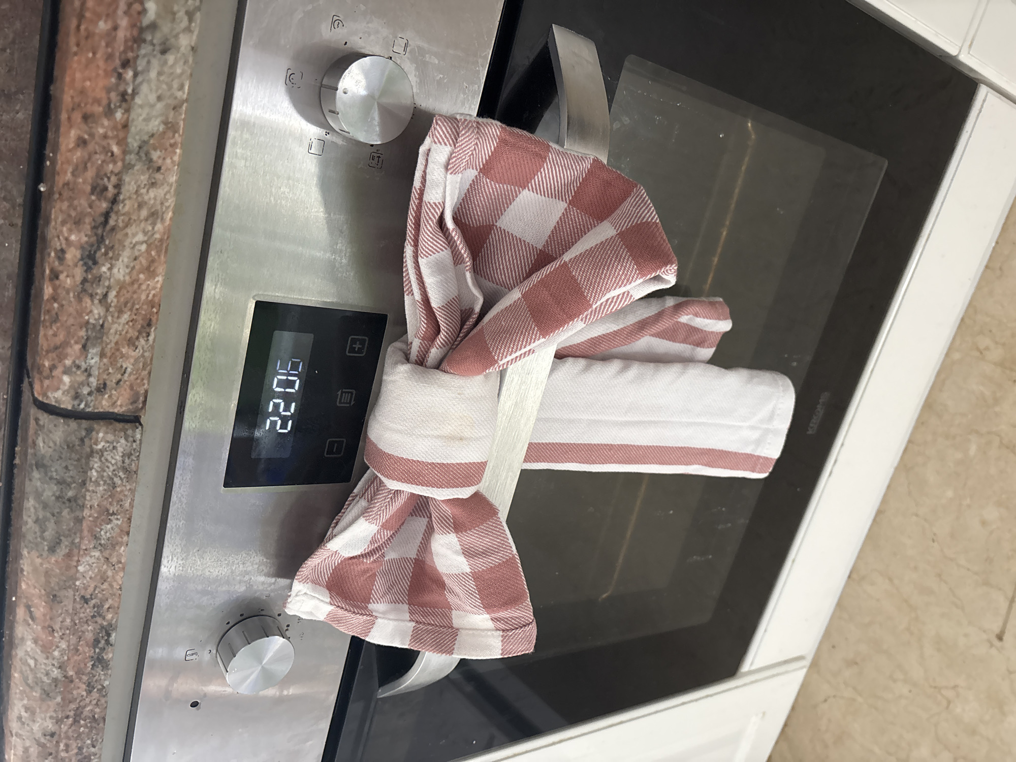 The image shows a close-up of a stainless steel oven with a digital display. The display shows "22 0". A checkered dish towel, red and white, is tied around the oven's handle. The towel is cinched in the middle, creating a bow-like shape. The oven is set against a kitchen counter with a granite-like pattern. The overall scene suggests a domestic setting, likely a kitchen in Orihuela, Spain. No people are visible, and the time of day and weather are not discernible. The image is well-lit, and there are no apparent emotions conveyed. The visible text on the oven's digital display reads "22 0".