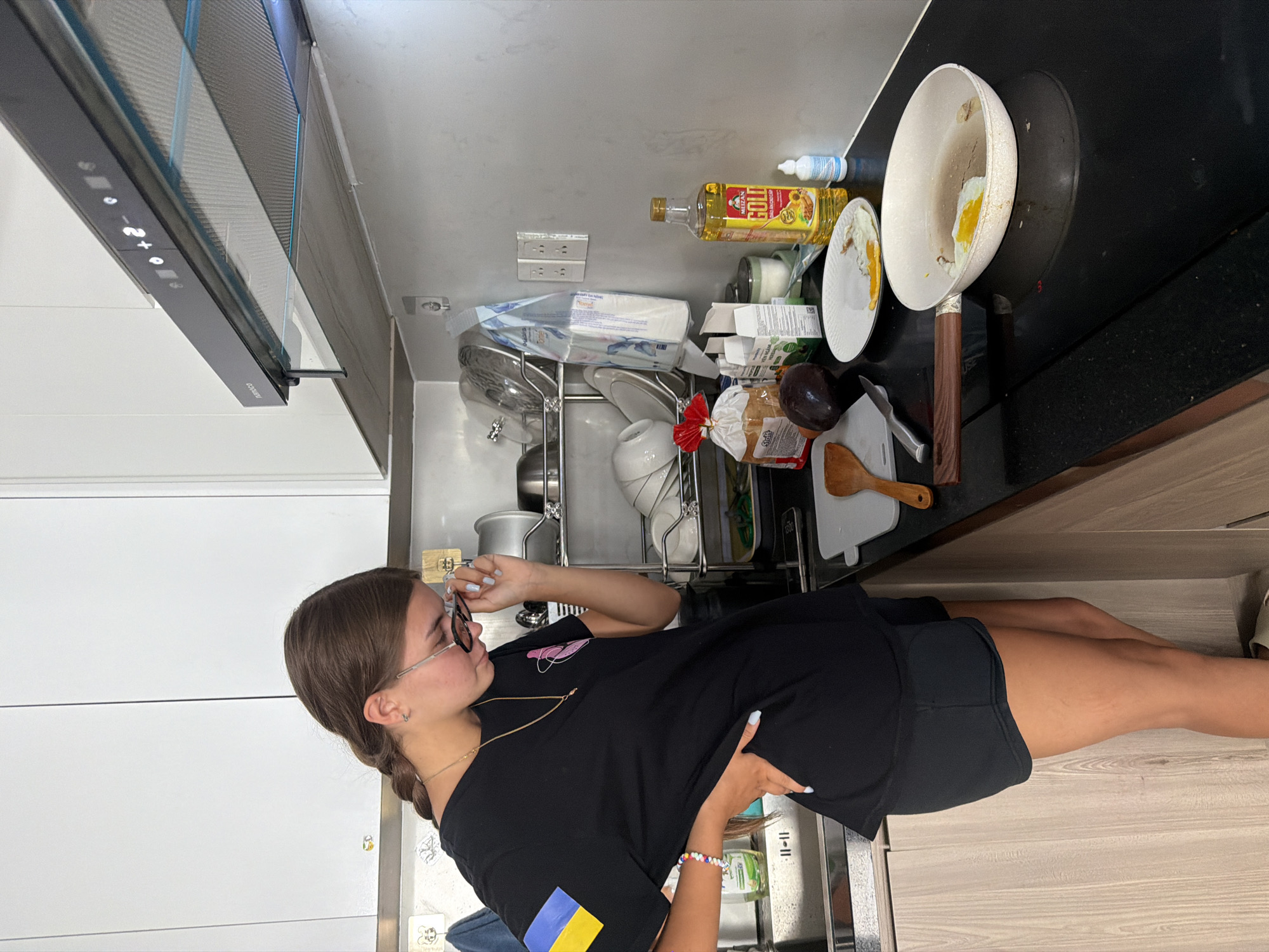 A young woman with brown hair in a braid, wearing a black t-shirt with a Ukrainian flag patch on the left sleeve and black shorts, stands in a kitchen, looking down at a cutting board. She is adjusting her glasses with her right hand, which is also resting on her hip. In front of her on the black countertop are a white plate with a fried egg, a frying pan with a fried egg, a loaf of bread in a red plastic bag, an avocado, a knife, a wooden spatula, a bottle of cooking oil, and a small white bottle. Behind her are white kitchen cabinets, a stainless steel sink, and a drying rack with dishes. The scene appears to be a domestic kitchen.