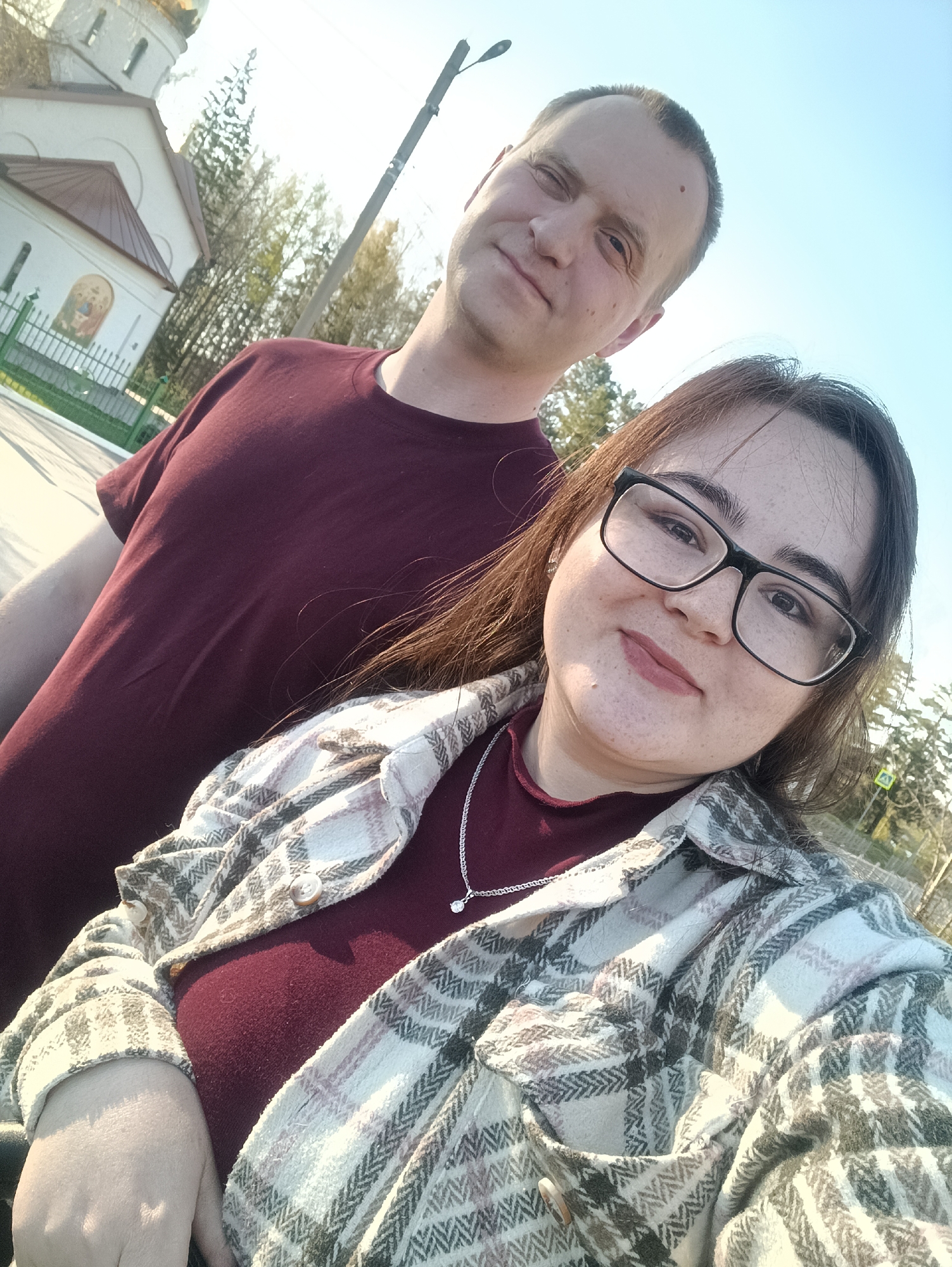 A selfie captures a man and a woman against a bright, daytime sky with a building in the background, potentially the city of Noviy, Russia. The man, positioned slightly behind the woman, wears a maroon t-shirt and has a neutral expression. The woman is in the foreground, wearing glasses and a plaid shirt over a maroon top, also displaying a necklace. The backdrop features a building with a dome, suggesting a church or religious structure, alongside trees and a street. A light pole is also visible. The overall setting implies an outdoor scene.