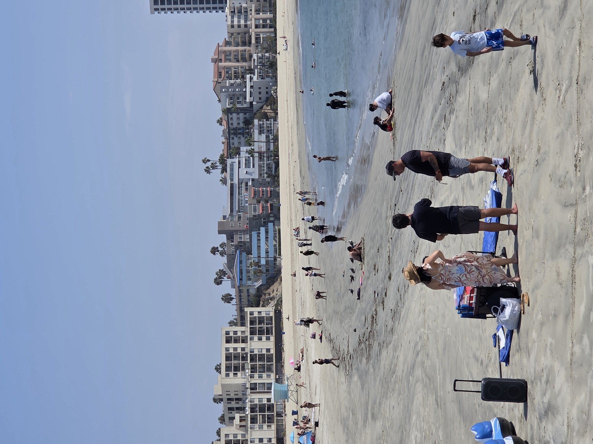 The image depicts a crowded beach scene on a sunny day in Long Beach, United States of America, with the Pacific Ocean in the background. In the foreground, several people are present. A woman in a floral dress and a straw hat stands with her back to the camera, conversing with a man in a black t-shirt and shorts. To their left, a large black speaker sits on the sand next to a rolling cart with beach items. Further to the right, a young boy in a blue t-shirt and patterned swim trunks walks across the sand. In the mid-ground, various individuals and groups are scattered across the beach and in the shallow water. Some are wading, while others are sitting or playing on the sand. A few seagulls are also visible near the water's edge. The background is dominated by a line of multi-story apartment buildings and hotels, with palm trees interspersed. The sky is clear and blue.