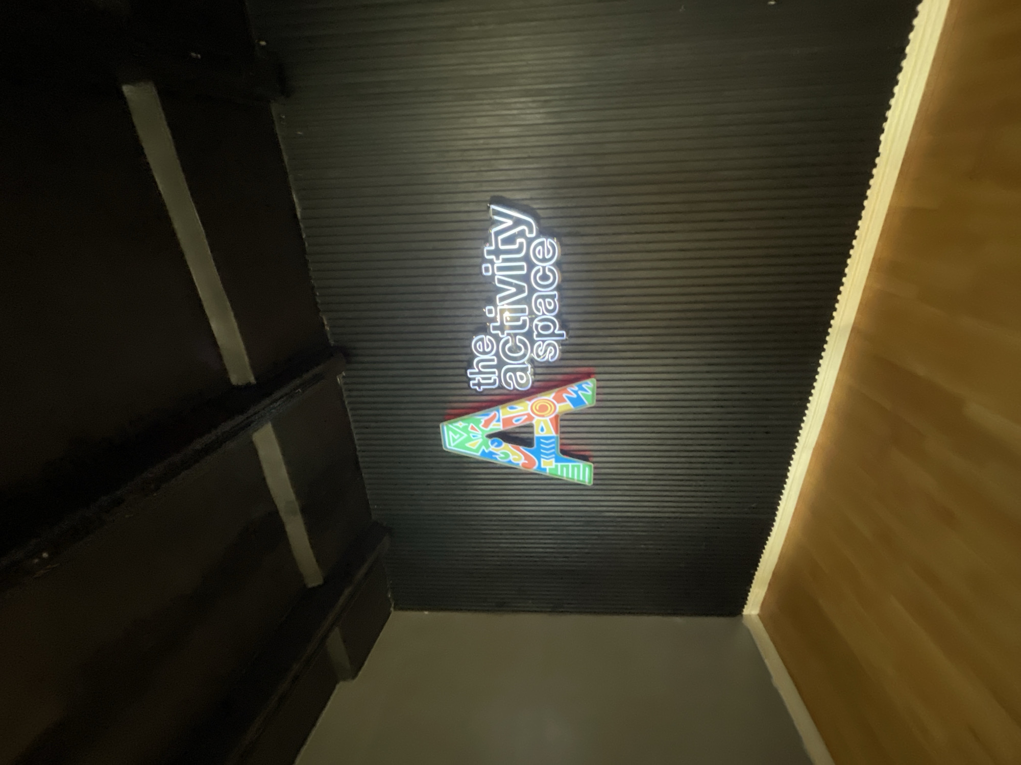 An indoor scene features a dark, ribbed ceiling with prominent signage. A large, stylized letter "A" is affixed to the ceiling, rendered in multiple bright colors and geometric patterns. Adjacent to the "A" is white neon lighting that spells out "the activity space." The perspective is from below, looking up diagonally.
To the left, a dark wall or structure with light-colored vertical elements extends from the foreground into the background, possibly indicating a staircase or a decorative panel. On the right, a section of light brown, possibly wooden, wall or paneling is visible, rising to meet the dark ceiling. The junction between the ceiling and this lighter wall has a distinct, jagged edge. The lower part of the frame reveals a plain, light grey surface, likely the floor or a lower wall section.
The space appears to be a modern commercial or recreational area, possibly an entrance or corridor, with the signage clearly indicating its purpose. There are no people visible, and no specific activity is taking place other than the static display of the architectural elements and signage. The lighting is ambient, with the neon sign providing a distinct glow against the dark ceiling. The overall impression is one of a designed indoor environment in Lekki, Nigeria.