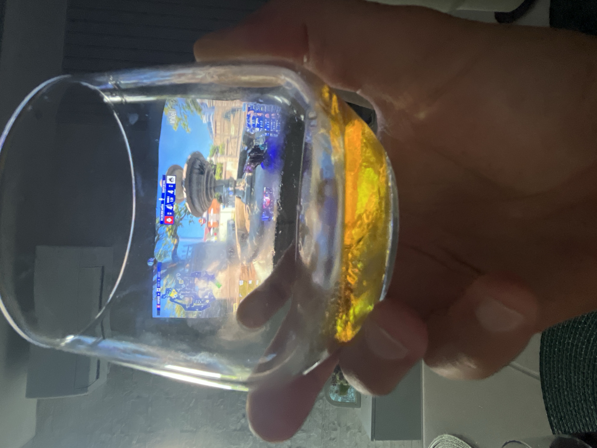 The image shows a person's hand holding a clear glass filled with a yellow liquid. The glass is positioned in front of a screen displaying what appears to be a video game. The game screen shows a colorful, outdoor scene with a fountain and buildings, suggesting a European-inspired environment. There are also some UI elements visible on the screen, including numbers and icons, typical of a game interface. The hand holding the glass is illuminated, and the liquid inside the glass reflects the light and the game screen, creating a warm, golden glow. The background is slightly blurred, but suggests an indoor setting. The time of day and weather are not discernible. The overall mood is casual and relaxed. Visible text on the screen includes "intel" and some game-related icons. The location context of Varna, Bulgaria is not directly evident from the image itself.