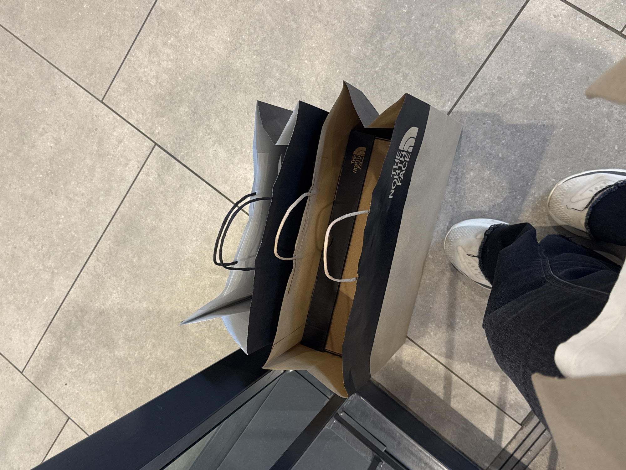 The image shows a view looking down at three shopping bags from The North Face. A person wearing dark-colored jeans and white sneakers is standing beside the bags. The bags are on a light gray tile floor, near a dark-colored window or frame. The location is likely an indoor space such as a store or shopping mall. There are no clues in the image to confirm that it is located in the city of Belgrade, Serbia.