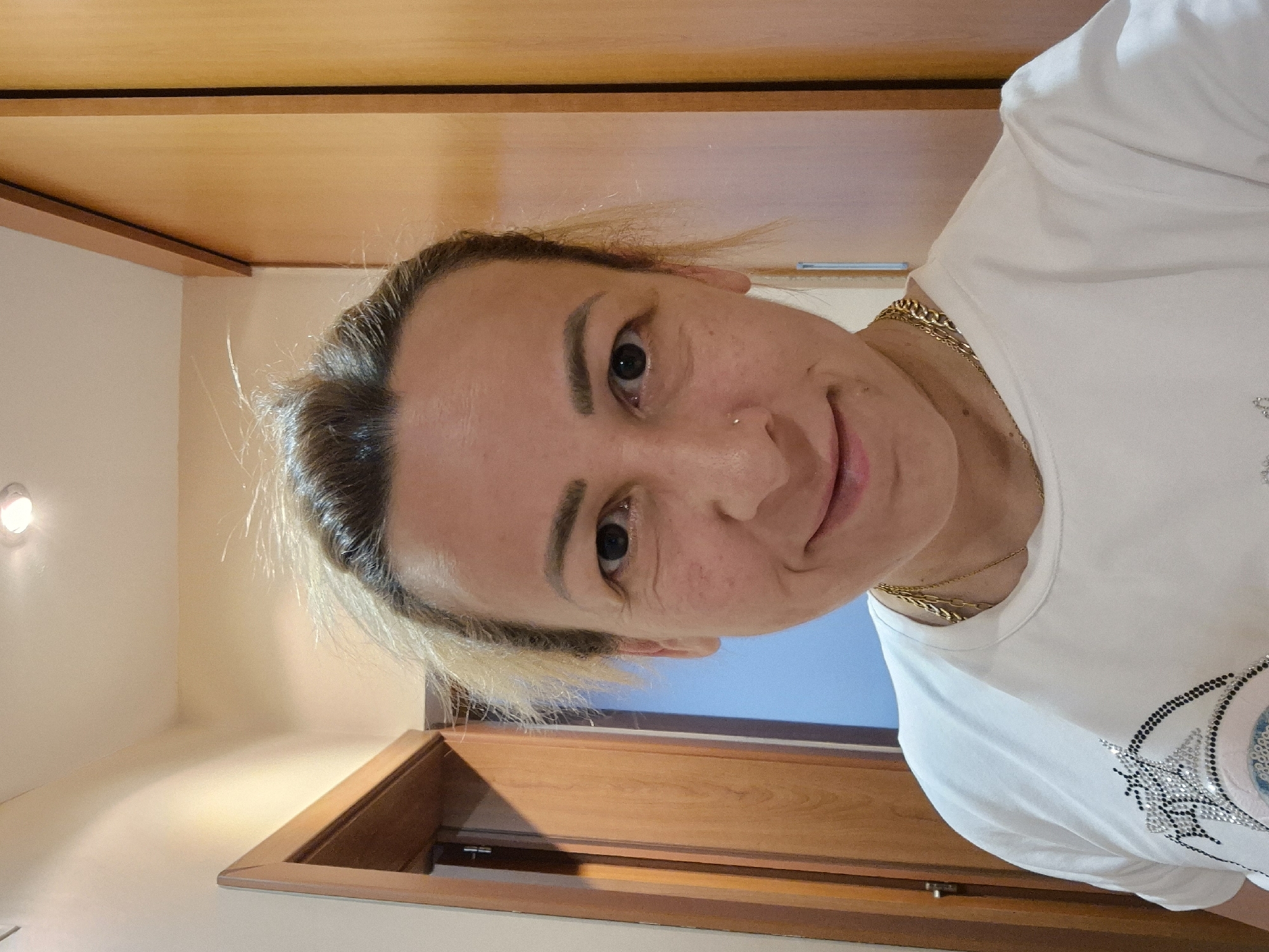 A medium shot captures a fair-skinned woman with blonde hair pulled back, standing in a hallway. She is wearing a white t-shirt with an embellished design on the front and a gold chain necklace. Her facial features include dark eyes, fair eyebrows, and a slight smile. She has a small nose ring. To her left is a closed wooden door, and to her right, a tall wooden cabinet. The hallway appears to be indoors, with a light fixture visible on the ceiling. The overall lighting is warm and natural.