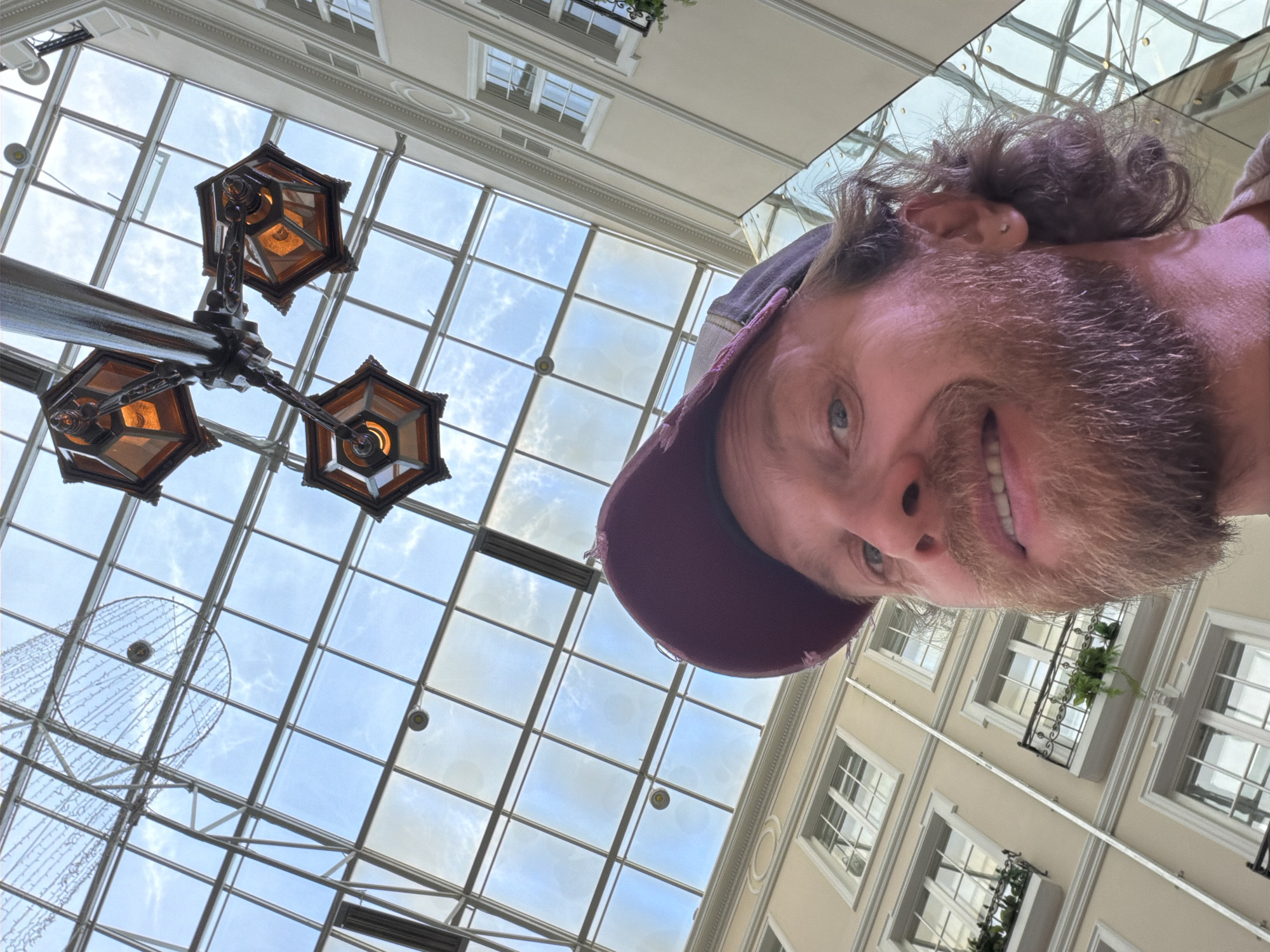 A man is looking up at a glass ceiling and holding his camera up with one hand to take a selfie. The man has a beard and is wearing a purple baseball cap. He is in the Passage Arcade in Saint Petersburg, Russia. The arcade has three ornate hanging lamps. The ceiling is supported by metal beams.