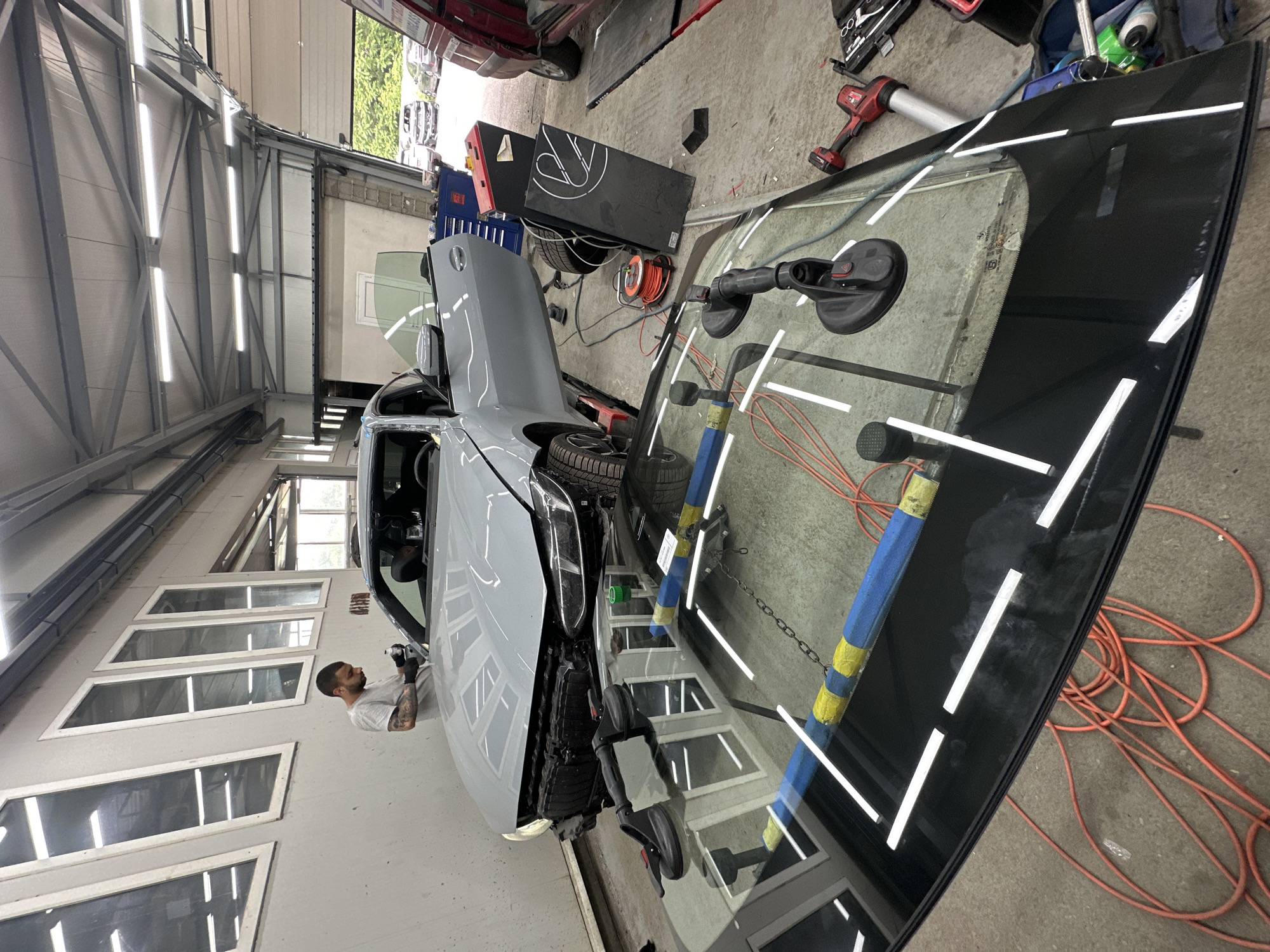 The image displays an indoor automotive workshop scene. A light-colored car, appearing to be a gray or light blue SUV, is positioned with its hood open on the left side of the frame, indicating ongoing repair or maintenance. A man with a beard and tattoos, wearing a white t-shirt and gloves, is actively working on the driver's side of the car, near the windshield frame or A-pillar, holding a tool.
In the foreground and to the right, a new car windshield stands upright, secured by two large black suction cups, suggesting it is being prepared for installation. The highly reflective surface of the windshield mirrors the bright, horizontal fluorescent lights lining the workshop's ceiling and walls, as well as reflections of the man and the interior of the workshop. A faint BMW logo is discernible on the concrete floor through the glass.
The workshop features an industrial design with metal framing, wall panels, and multiple rectangular windows on a side wall. The concrete floor is scattered with various tools and equipment, including a red power drill, entangled orange electrical extension cords, car tires, and two black boxes with white outlines, which could be specialized equipment like heating lamps or calibration tools. The scene is brightly illuminated by artificial lighting, characteristic of a professional vehicle service center, likely undertaking windshield replacement or bodywork. No specific time of day or weather is indicated due to the indoor setting.