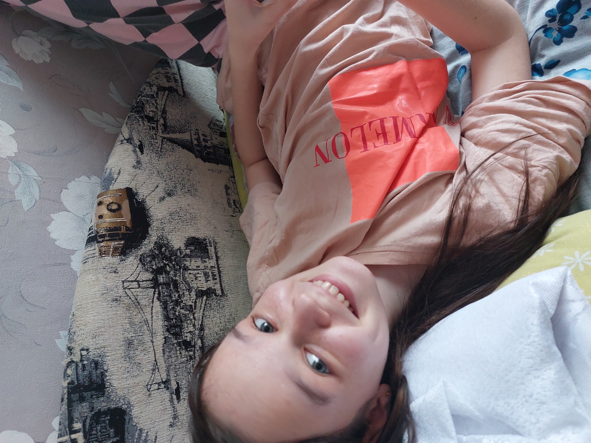 A young person with fair skin, blue eyes, and dark hair is lying on what appears to be a patterned sofa or bed. They are wearing a peach-colored t-shirt with the word "MELON" in red print on a coral-colored graphic. Their body is angled, and they are smiling at the camera. The background includes part of a pink and black checkered pattern, floral wallpaper, and a patterned fabric. No specific details confirm this location as Большой Букор, Russia.