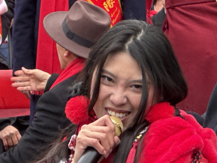 The image captures a close-up of a vibrant public event, prominently featuring a woman performing or speaking, surrounded by a festive atmosphere.
In the foreground, a woman with long, dark hair is intensely focused, holding a gold-colored microphone with her right hand. Her facial expression is animated and energetic, characterized by a wide, open-mouthed smile revealing her teeth, and crinkled eyes that convey passion or excitement. Her dark hair is styled with a center part and falls over her shoulders. She is dressed in red attire, which appears to be fuzzy or made of a soft material, and features large red pom-poms visible on her shoulders or attached to her clothing. Red tassels are also visible as part of her outfit.
Behind the main subject, to the upper left (our right), another individual is partially visible from the back. This person is wearing a dark brown fedora-style hat with a black band and a prominent red scarf draped around their neck. Their left hand is raised in front of them, palm open, suggesting a gesture, perhaps clapping or signaling. A dark-colored jacket is also visible on this individual.
The background further suggests a crowded setting. Red fabric, likely part of clothing or decorations, is visible throughout the frame, including what appears to be a red padded seat or bench in the mid-ground. In the upper left portion of the image, a red banner or sign is partially visible. On this banner, the English text "Grand Marshal" can be clearly read, although some characters above it are obscured and indecipherable. The dominance of red colors, the performance aspect from the microphone, and the "Grand Marshal" text collectively suggest a parade, celebration, or a significant public gathering. The overall mood conveyed is one of excitement and engagement.