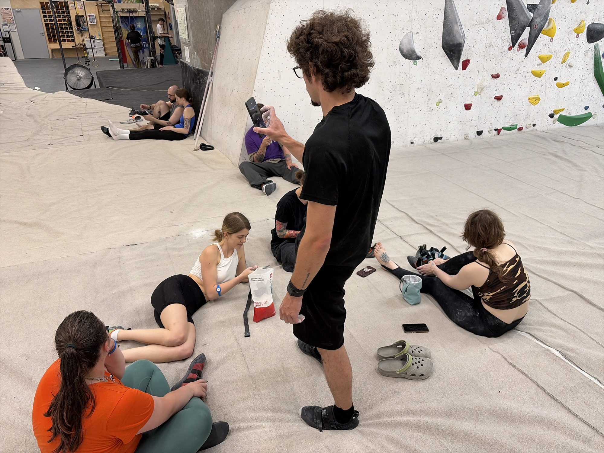 In a climbing gym in Khimki, Russia, several people are gathered on the floor. A man in a black t-shirt and shorts stands in the foreground, looking at his phone. Behind him, a person in a purple shirt sits with their legs crossed. To the left, two individuals, a man and a woman, are seated, with the woman wearing a blue tank top and the man in a dark shirt. In the middle ground, a woman in a white tank top and black shorts is lying on the floor, reaching for a bag of chalk. To her right, a person in a camouflage tank top and black leggings is seated. In the lower left, a person in an orange t-shirt and green leggings sits with their back to the camera. The environment is a climbing gym with padded flooring and a climbing wall visible in the background.