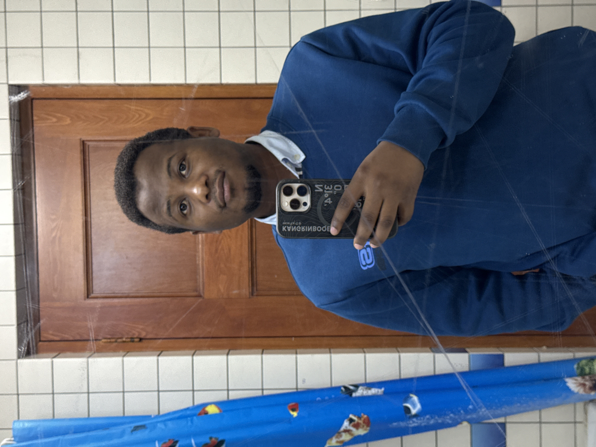 A man is taking a selfie in a bathroom mirror. The man is wearing a blue sweater and a collared shirt. The bathroom has tiled walls and a brown wooden door. A blue shower curtain is visible in the lower part of the frame. There is no information about the location of the image, and no information suggests this image was taken in the city of Daxing District, China.