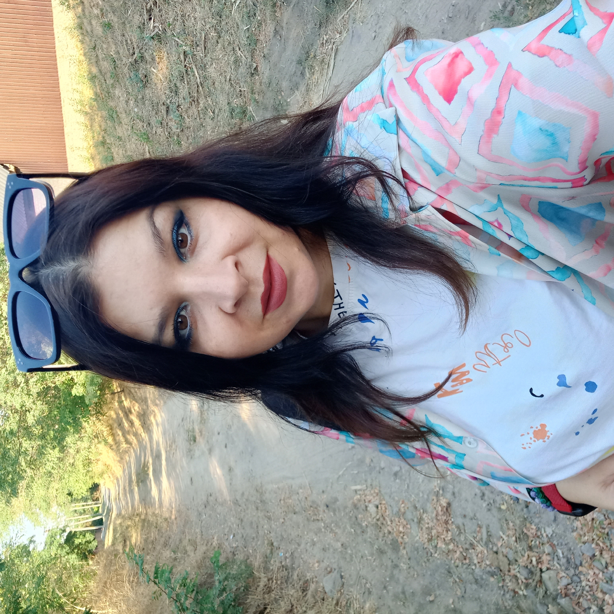 A woman with long, dark hair and dark sunglasses perched on her head takes a selfie outdoors. She is wearing a white t-shirt with colorful abstract designs and a light-colored, patterned open shirt. Her makeup includes blue eyeshadow and dark lipstick. She is standing on a dirt path, with dry grass and trees on either side. In the background, a brown wooden building is visible. The location is a rural area outside of Klyntsi, Ukraine.