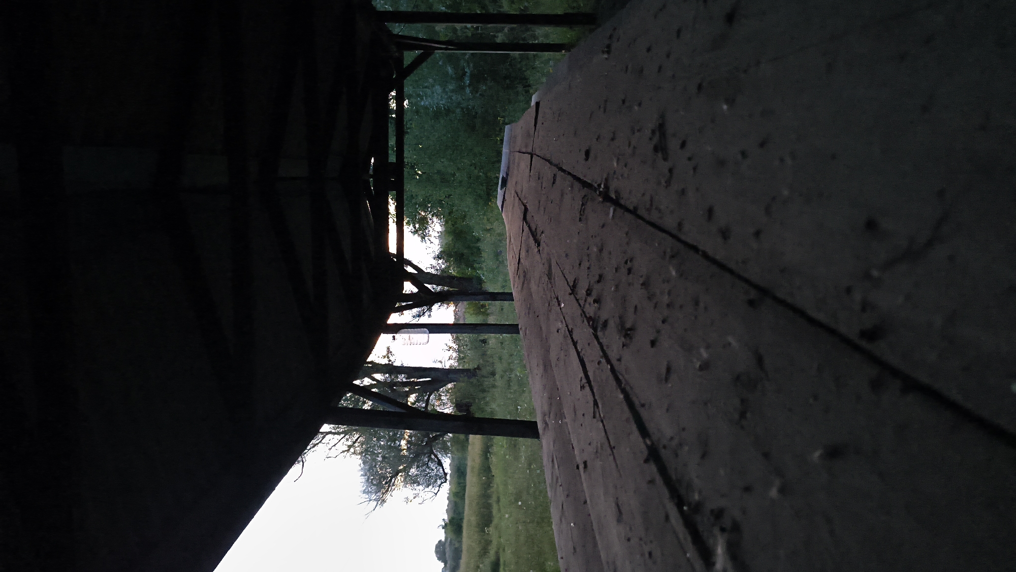 A low-angle, full shot shows a weathered, rustic picnic table under a dark wooden pavilion. The table's surface is rough and covered in dust and debris, with visible cracks running along its length. The pavilion's roof is dark and shadowed, obscuring the structure above. Through the open sides of the pavilion, a grassy field stretches into the distance, dotted with trees and low vegetation. The sky is a pale, hazy white, suggesting either early morning or late evening. The scene is tranquil and untouched, devoid of any human presence.