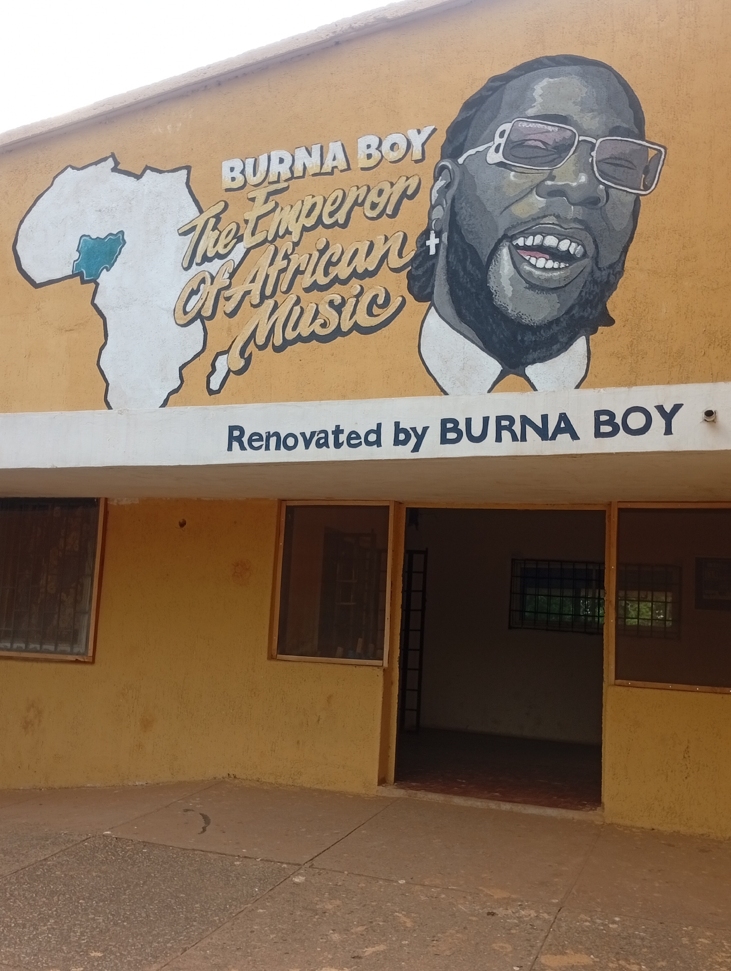 This is a medium shot of a mural painted on the exterior wall of a building. The mural features a portrait of Burna Boy, a Nigerian musician, wearing glasses and a white shirt. To the left of his portrait, there is a stylized outline of the continent of Africa, with Nigeria highlighted in a blue color. Text above Burna Boy's portrait reads "BURNA BOY The Emperor Of African Music". Below the mural, a white band across the building has the text "Renovated by BURNA BOY" painted in dark blue letters. The building has a yellow facade, and there are several windows and an open doorway visible. The ground in front of the building is paved with concrete tiles. The scene appears to be outdoors, during daylight. The overall impression is one of commemoration and artistic expression.