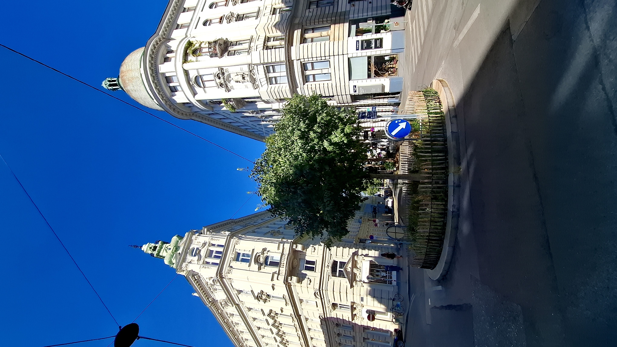 The image captures a sunny day in a city, likely Vienna, Austria, characterized by ornate architecture and clear blue skies. In the foreground, a circular tree planter is positioned in the center of a street intersection. The planter is surrounded by a low wooden fence and contains a medium-sized tree with dense green foliage. A blue traffic sign with a white arrow pointing to the right is visible within the planter.
Flanking the intersection are two distinct, multi-story buildings. The building on the left features a pointed green spire with a clock face and ornate decorative elements on its cream-colored facade, typical of Viennese architecture. The building on the right has a rounded corner and a green dome, also adorned with classical architectural details. Both buildings appear to be commercial establishments on the ground floor, with windows and balconies on the upper levels.
The street is paved, with visible road markings and a crosswalk. Several bicycles are parked near the building on the left. Two individuals are walking on the sidewalk in front of the building on the left. The overall impression is a vibrant urban scene during daylight hours.