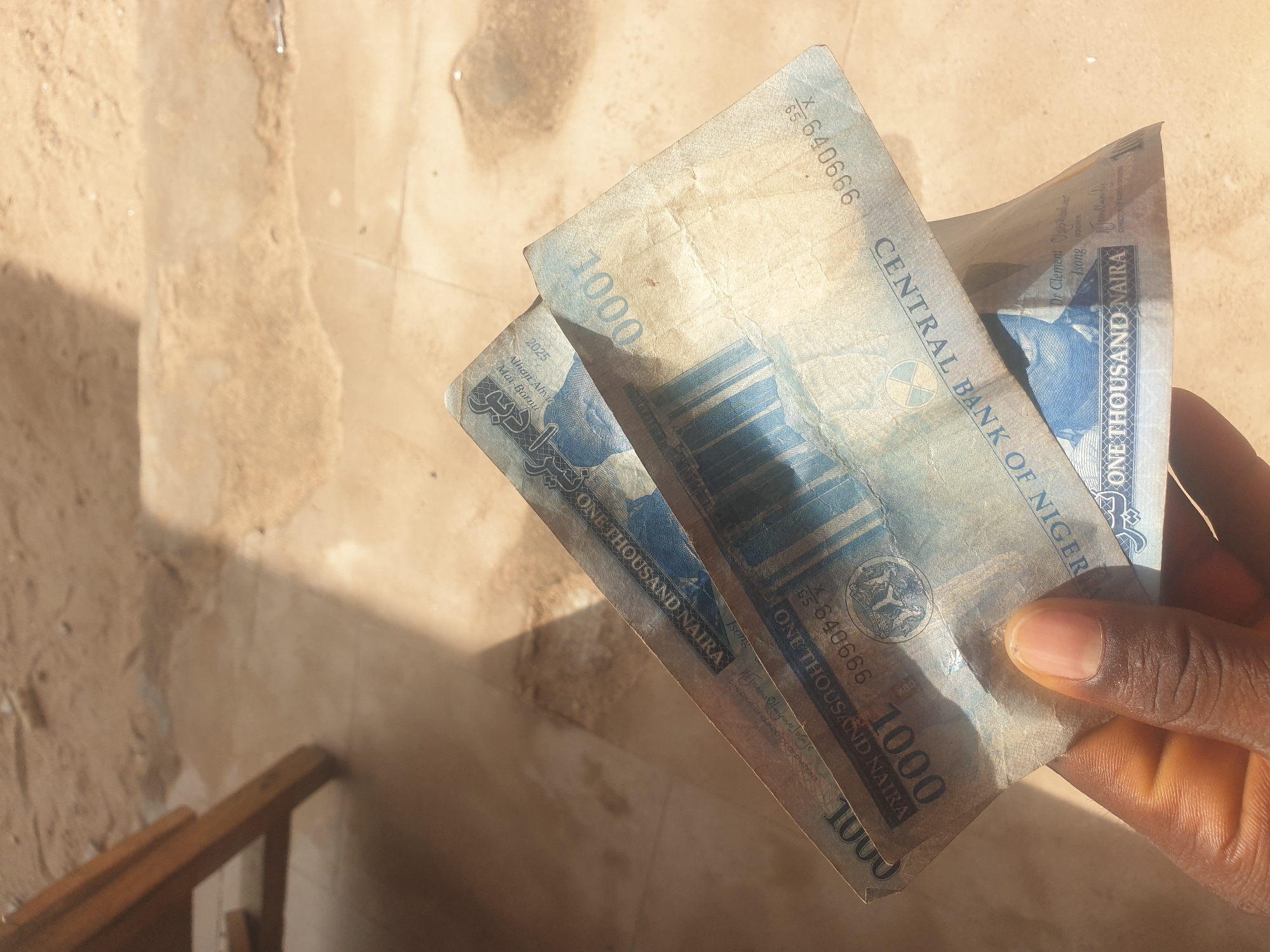 A person's hand, with a dark complexion, holds two Nigerian 1000 Naira banknotes. The banknotes are oriented diagonally, with the top one partially obscuring the bottom one. The banknotes appear worn and slightly creased. The background consists of a textured, sandy surface with patches of light and shadow. The image is taken outdoors, as suggested by the natural lighting and the sandy ground. The context of Jauro Yinu, Nigeria, implies this is likely a street scene or an outdoor market area.
