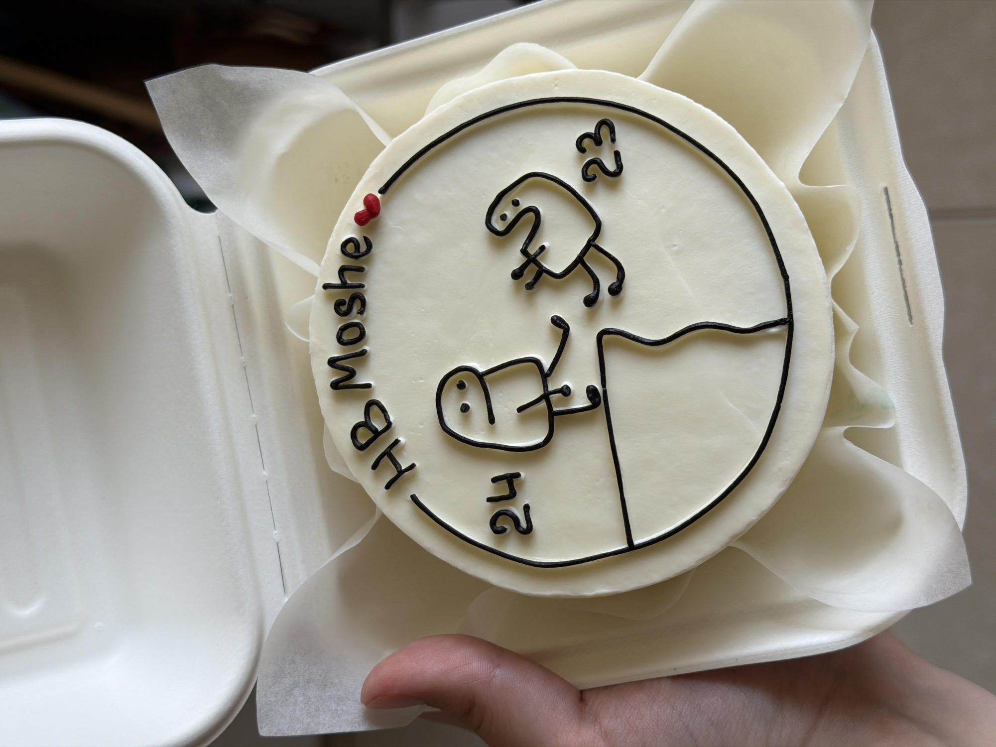 A small, personalized celebration cake, predominantly white and circular, is held within a disposable take-out container. The cake is nestled on white, translucent parchment paper that lines the container. A hand, with fingers visible from below, supports the container.
The cake's top surface features a minimalist design piped in black icing. A large black circle frames the central imagery. Inside this circle, two stylized, blob-like figures with simple faces and stick limbs are depicted. One figure stands on a higher ledge, with the number "23" piped beside it. Below, another figure appears to be falling or climbing, clinging to a cliff-like edge, with the number "24" adjacent.
Curving along the left edge of the cake, outside the main circle, the text "HB Moshe" is written in black icing, signifying "Happy Birthday Moshe." A small, bright red heart is placed above the "HB" part of the text, adding a touch of color and affection. The cake's white base contrasts sharply with the bold black outlines and text, making the design stand out. The scene suggests a lighthearted and personal celebration. No specific time of day or external location cues are visible beyond the immediate context of the cake being held in its packaging. The provided context indicates the scene takes place in Tel-Aviv, Israel.