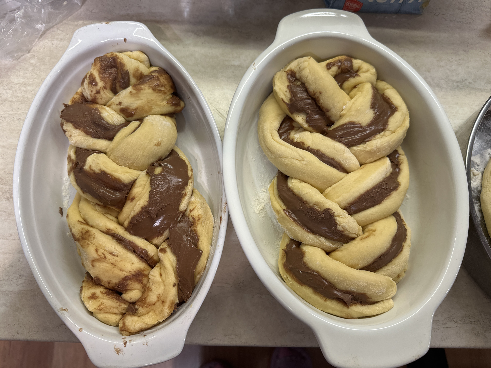 Two braided loaves of dough with chocolate filling are in white oval dishes on a countertop. The loaves appear to be ready for baking. The left loaf has a darker, more spread-out chocolate filling. The right loaf's filling is more contained. A bag and a container with flour are visible in the background. A pan with raw dough is on the right of the image. The location is unknown, therefore I cannot provide an accurate depiction of Sofia, Bulgaria.