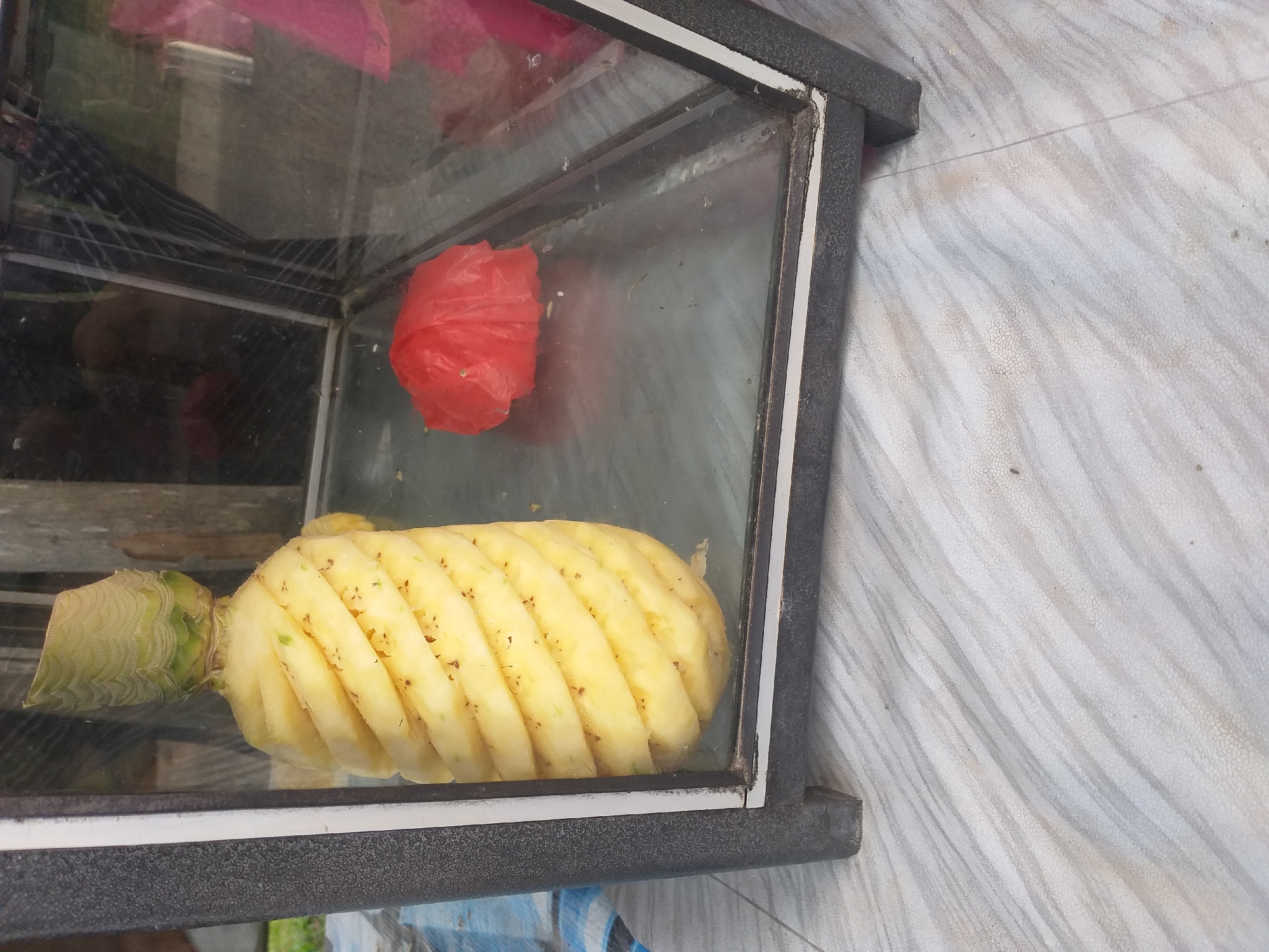 In Air Jamban, Indonesia, a partially peeled pineapple stands upright inside a glass display case. The pineapple's skin has been spirally removed, revealing the yellow flesh. The display case is framed by a dark-colored material. Inside the case, to the right of the pineapple, a crumpled red plastic bag is visible. The display case rests on a textured surface with a patterned design. The background of the image is blurred, but appears to be outdoors with some vegetation.