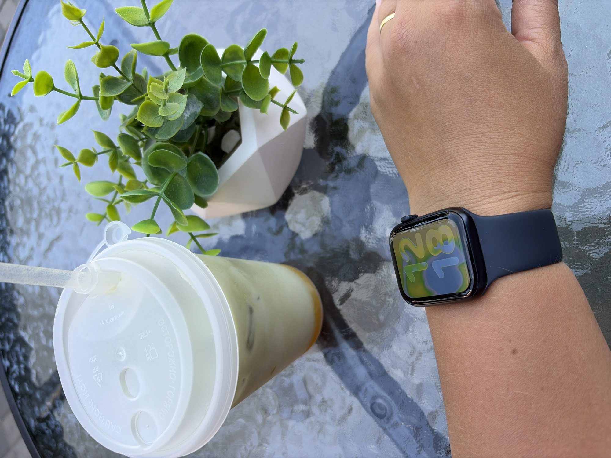 An individual's left wrist, adorned with a black smartwatch displaying the time 12:18 with a colorful watch face, rests on a reflective glass table. Next to the wrist is a clear plastic cup filled with a pale green beverage and ice, topped with a white lid and a clear plastic straw. A small potted plant with green leaves sits to the right of the cup. The scene is set outdoors, likely on a table in a city park or cafe, given the reflective glass surface and the presence of a beverage and decorative plant. The image was taken on a table in the city of Kommunarka, Russia.