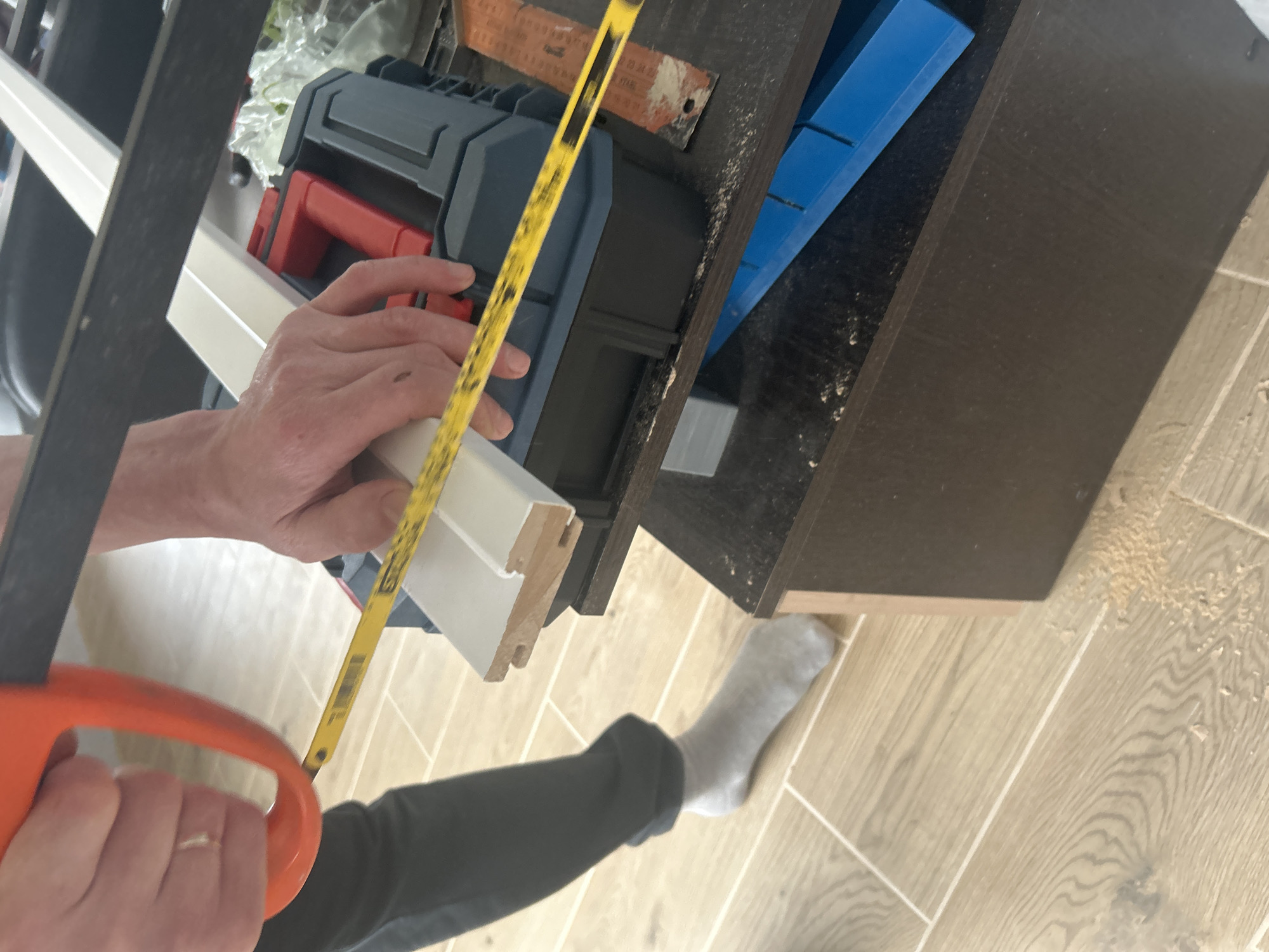 A person wearing dark-colored pants and a gray sock is cutting a white molding using a hand saw with an orange handle. The person holds a yellow tape measure against the molding. The molding is positioned against a dark-colored cabinet with a blue miter box. A toolbox is sitting on top of the cabinet. The floor is covered with light-colored wood-patterned tiles. The scene appears to be indoors, possibly in a workshop or home improvement setting. The city of Chaikovsky, Russia is not visually evident in the image.