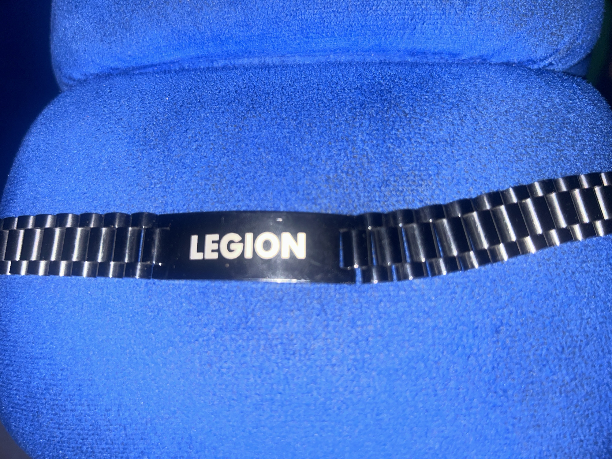 A close-up, vertically oriented photograph displays a metallic watchband with the word "LEGION" embossed in white on a black rectangular plate. The watchband is positioned on a textured blue surface, possibly fabric or upholstery. The image is grainy and shows some glare on the metallic surfaces. No people, specific locations in Ogbomosho, Nigeria, or any actions or interactions are visible.