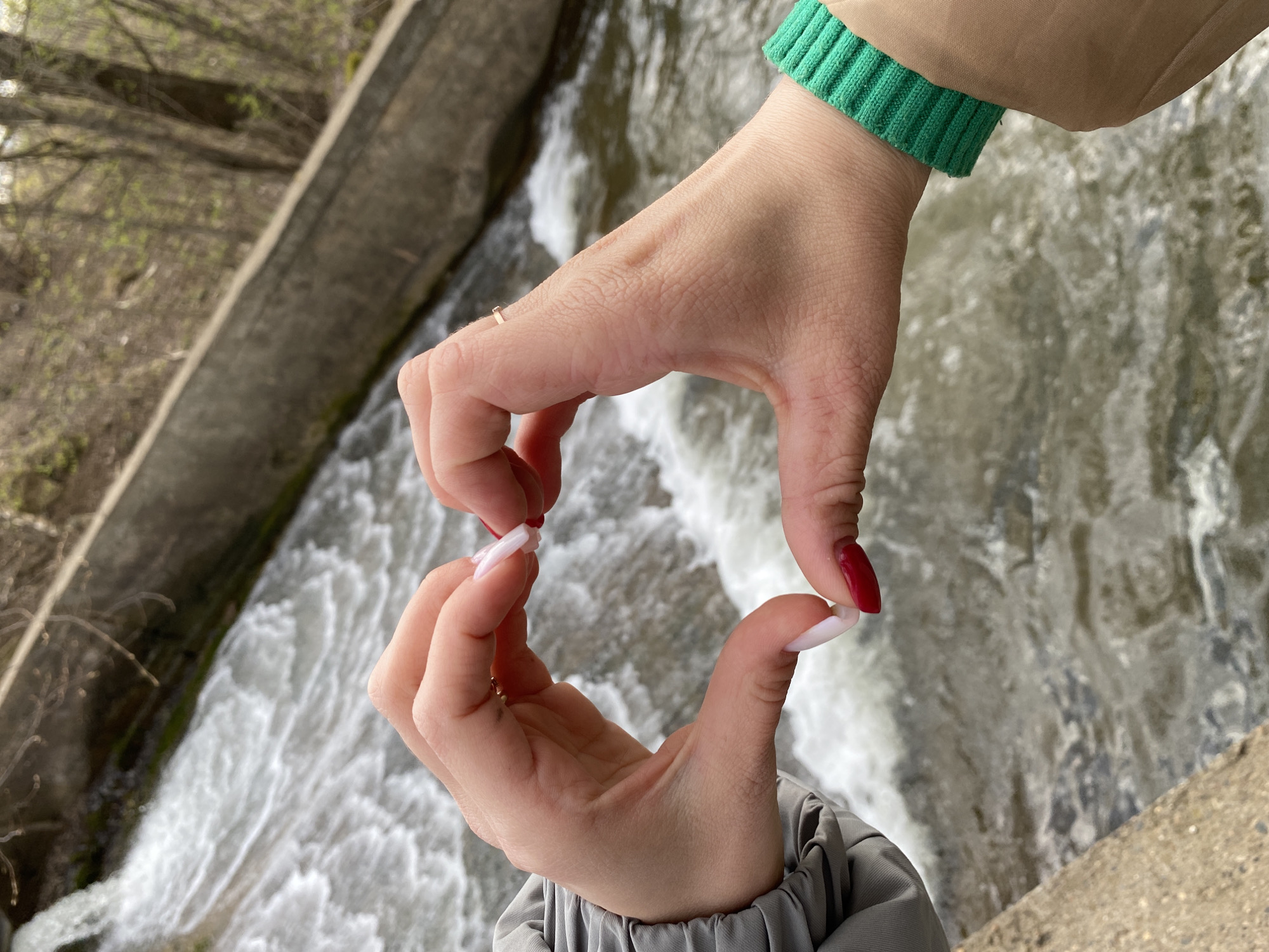 The image presents a top-down view of two hands forming a heart shape against a natural backdrop. The top hand is adorned with a green ribbed cuff, visible at the wrist of a beige jacket sleeve, and wears a small, gold-toned ring on its thumb. Its fingernails are natural or painted a very light, almost white, shade. The bottom hand, emerging from a grey, pleated sleeve, features striking red-painted fingernails. Both sets of nails are well-maintained, with the red ones appearing almond-shaped and the lighter ones having a squarer, longer tip.
The hands are positioned over a turbulent body of water, likely a river or stream, characterized by foamy white rapids and strong currents. Concrete structures, possibly part of a weir or dam spillway, line the banks of the waterway, visible along the left and bottom right edges of the frame. In the upper left background, bare tree branches with emerging buds or sparse foliage suggest the season is either early spring or late autumn. The lighting is bright and even, indicating it is daytime, possibly under an overcast sky, as no harsh shadows are cast. The heart gesture conveys a sense of love, appreciation, or connection with the natural environment. No text is visible in the image. The scene is set in Фоки, Russia.