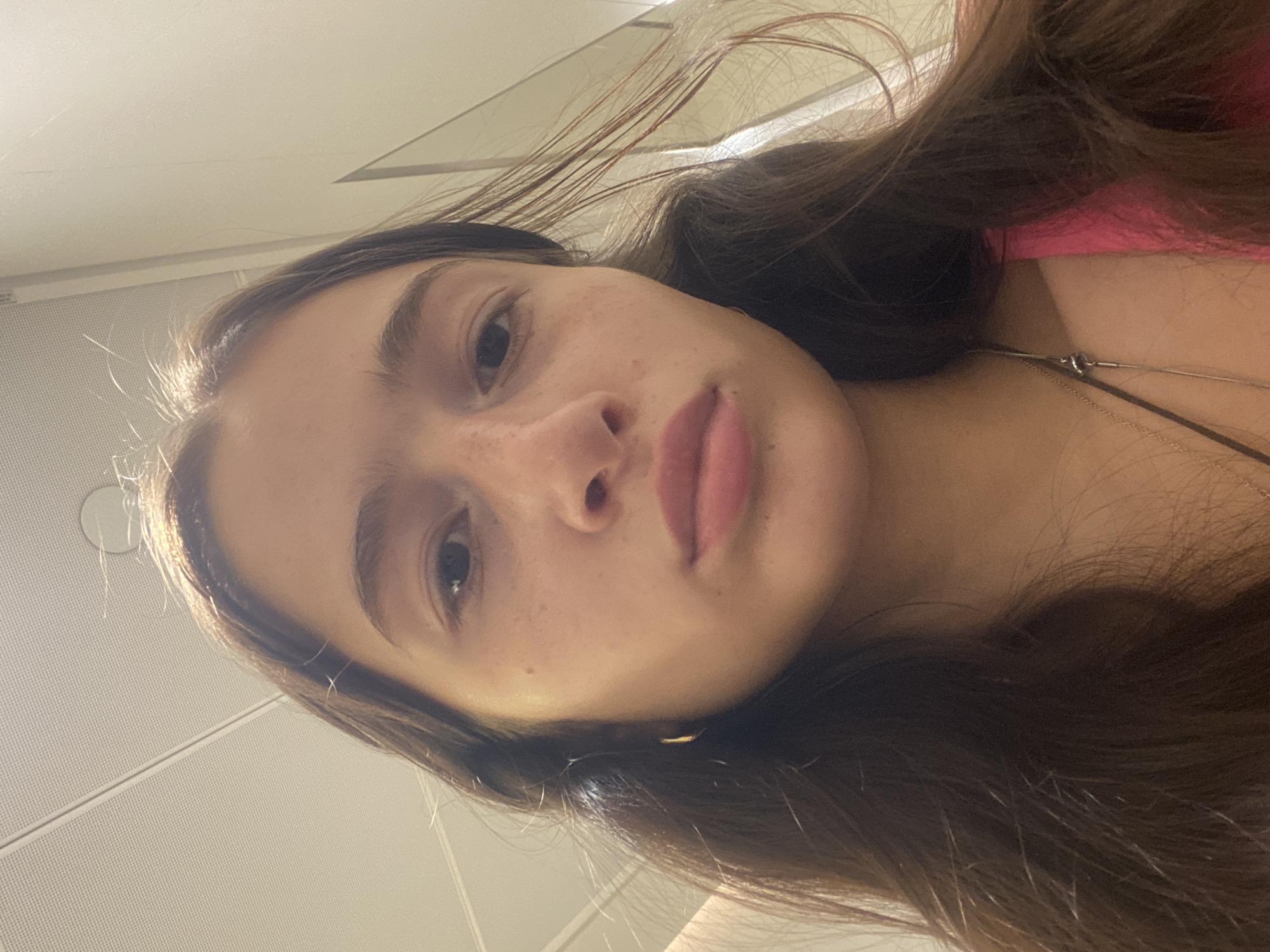 A close-up selfie captures a young woman with fair skin, brown hair, and freckles on her face. She is wearing a pink top and a silver necklace. The background appears to be the interior of a building with a white, perforated ceiling. There is no clear indication that the image was taken in Dubai, United Arab Emirates.