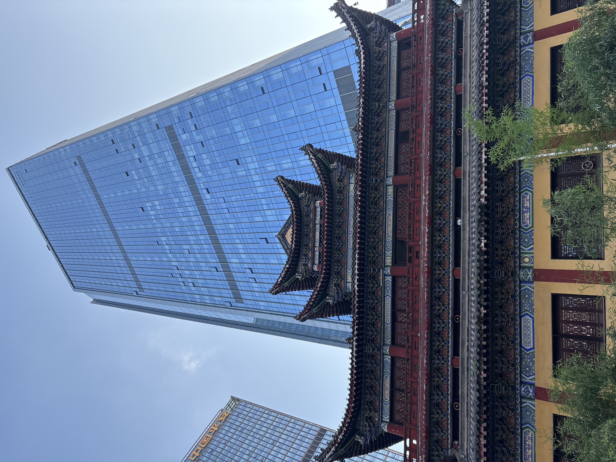 In the Yuzhong District of China, a traditional Chinese temple building with ornate red and blue decorative elements and tiered, curved roofs stands in the foreground. Behind the temple, two modern glass skyscrapers rise into a clear blue sky. One skyscraper on the left displays Chinese characters on its facade. Green bamboo plants are visible in front of the temple.