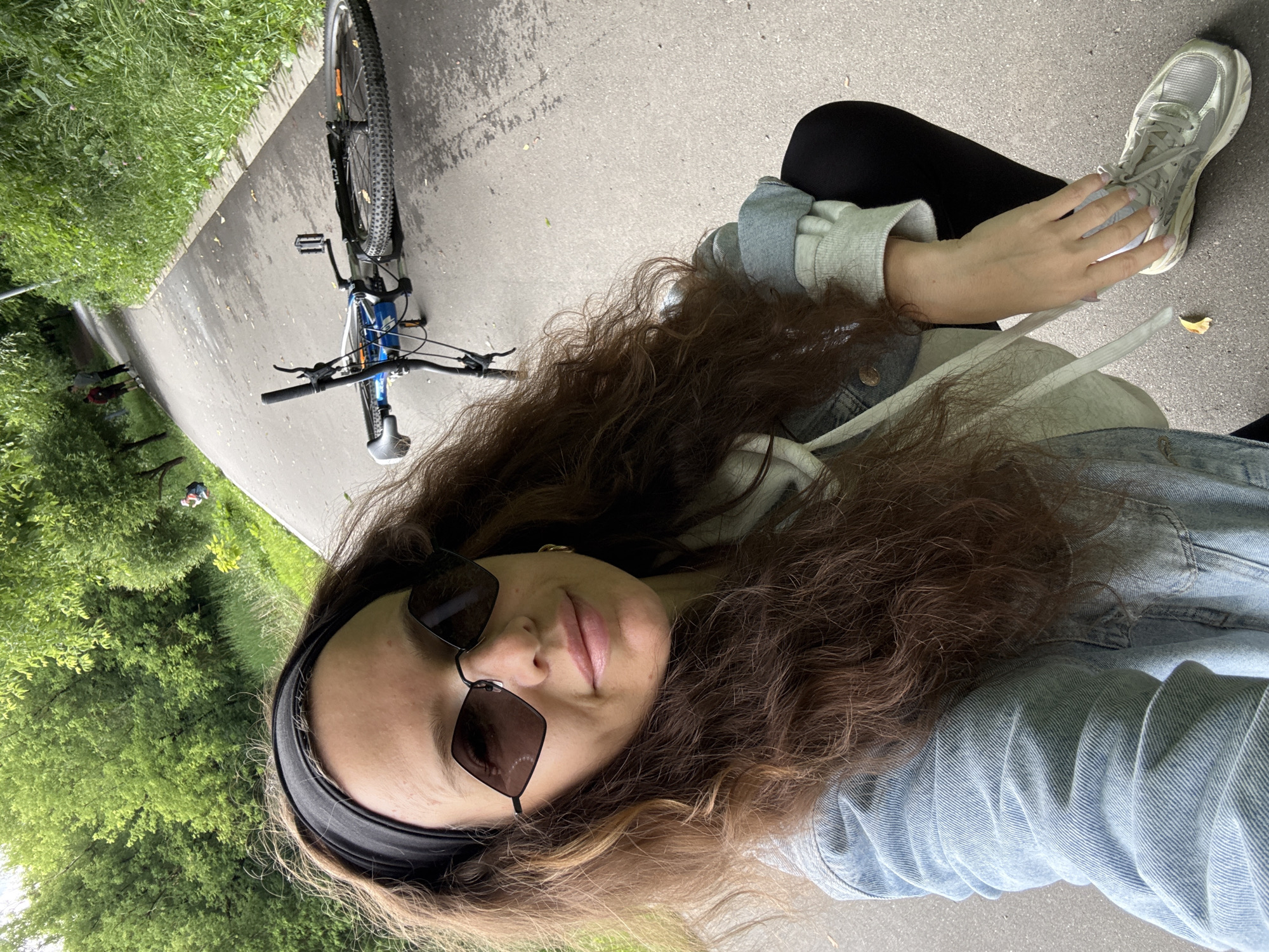 In Moscow, Russia, a woman with long, curly brown hair and sunglasses is kneeling on a paved path in a park. She is wearing a light blue denim jacket over a grey hoodie and black leggings. Her left hand is on her right shoe, tying the laces. A blue bicycle is lying on its side on the path behind her, its wheels facing away from the camera. The path is wet and appears to have recently rained. Lush green trees and grass line the path, with other people visible in the distance walking and cycling. The overall scene depicts a casual moment in an outdoor urban park.