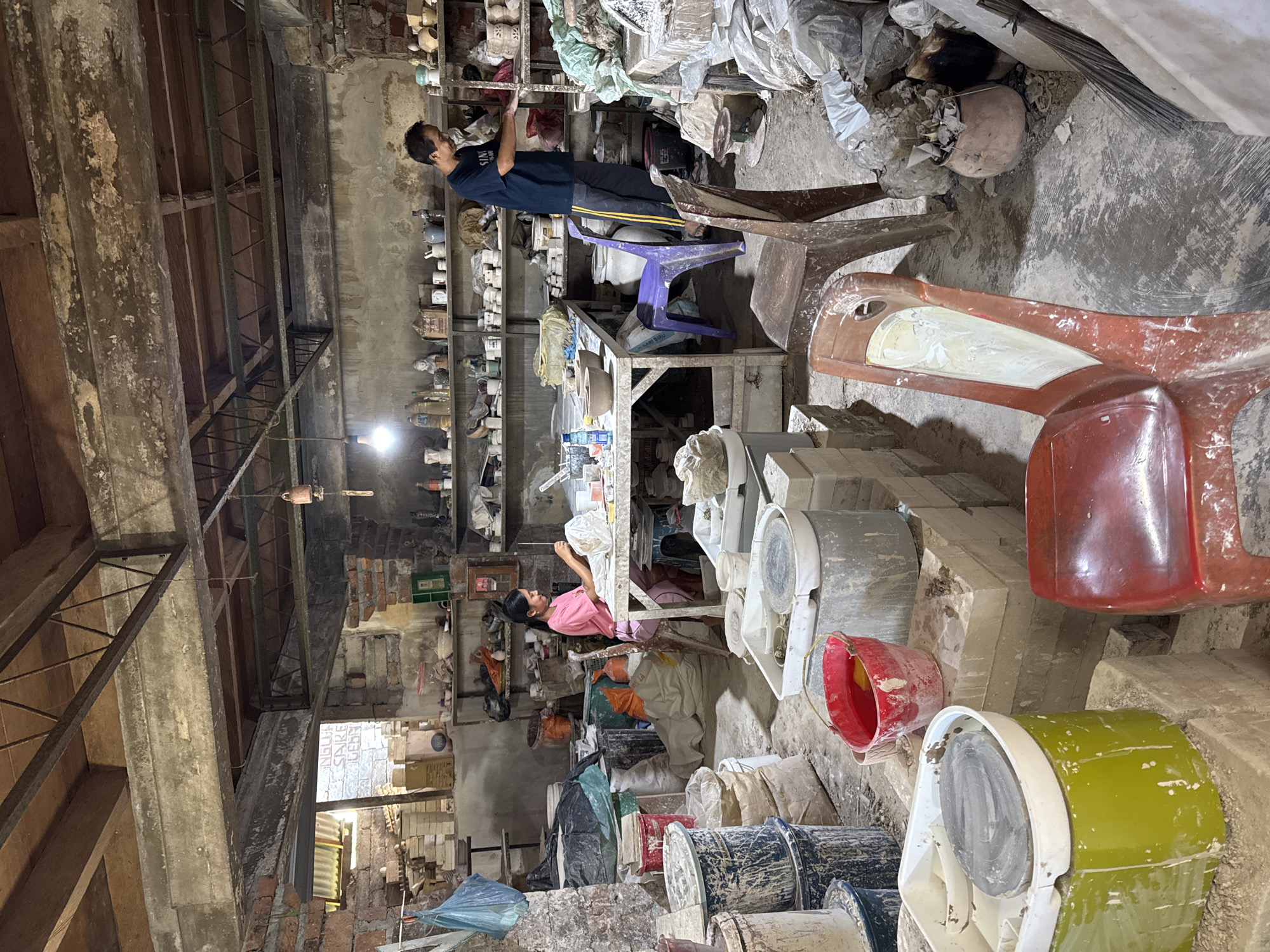 The photo shows the interior of a ceramic workshop located in Kediri, Indonesia. The workspace is cluttered and dusty. Two individuals are present: an individual wearing a blue shirt and black pants is standing and reaching towards a high shelf filled with ceramic molds. Another individual, wearing a pink shirt, is working at a table, possibly cleaning or assembling the ceramic pieces.
The environment consists of stacks of ceramic molds and completed pieces in various sizes and colors. Numerous paint buckets, both empty and partially filled, are scattered around the floor. The walls are made of aged brick and concrete. Overhead, there are exposed wooden beams supporting the structure. A hanging light fixture provides illumination. Various chairs are present, and the scene suggests ongoing ceramic production activity.