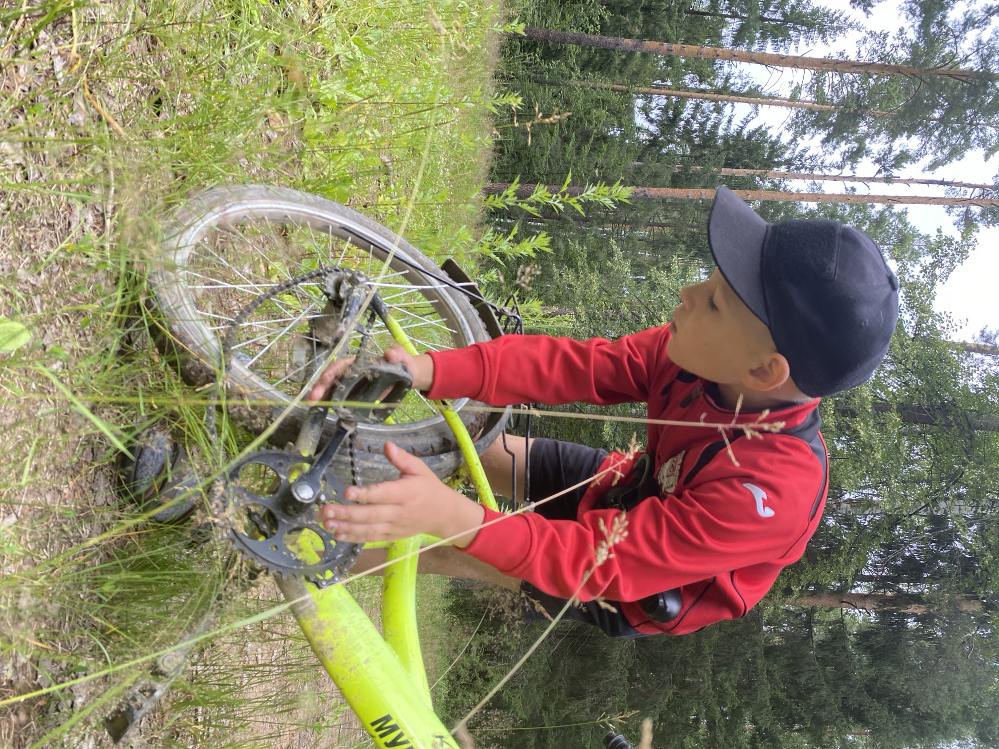 A young boy wearing a black baseball cap and a red long-sleeved shirt is in a forest in Noviy, Russia. He is bending over a bright yellow bicycle, with his hands on the gears and chain. The bicycle appears to be stuck or have a problem. The ground is covered in grass and dirt, and there are tall trees in the background.