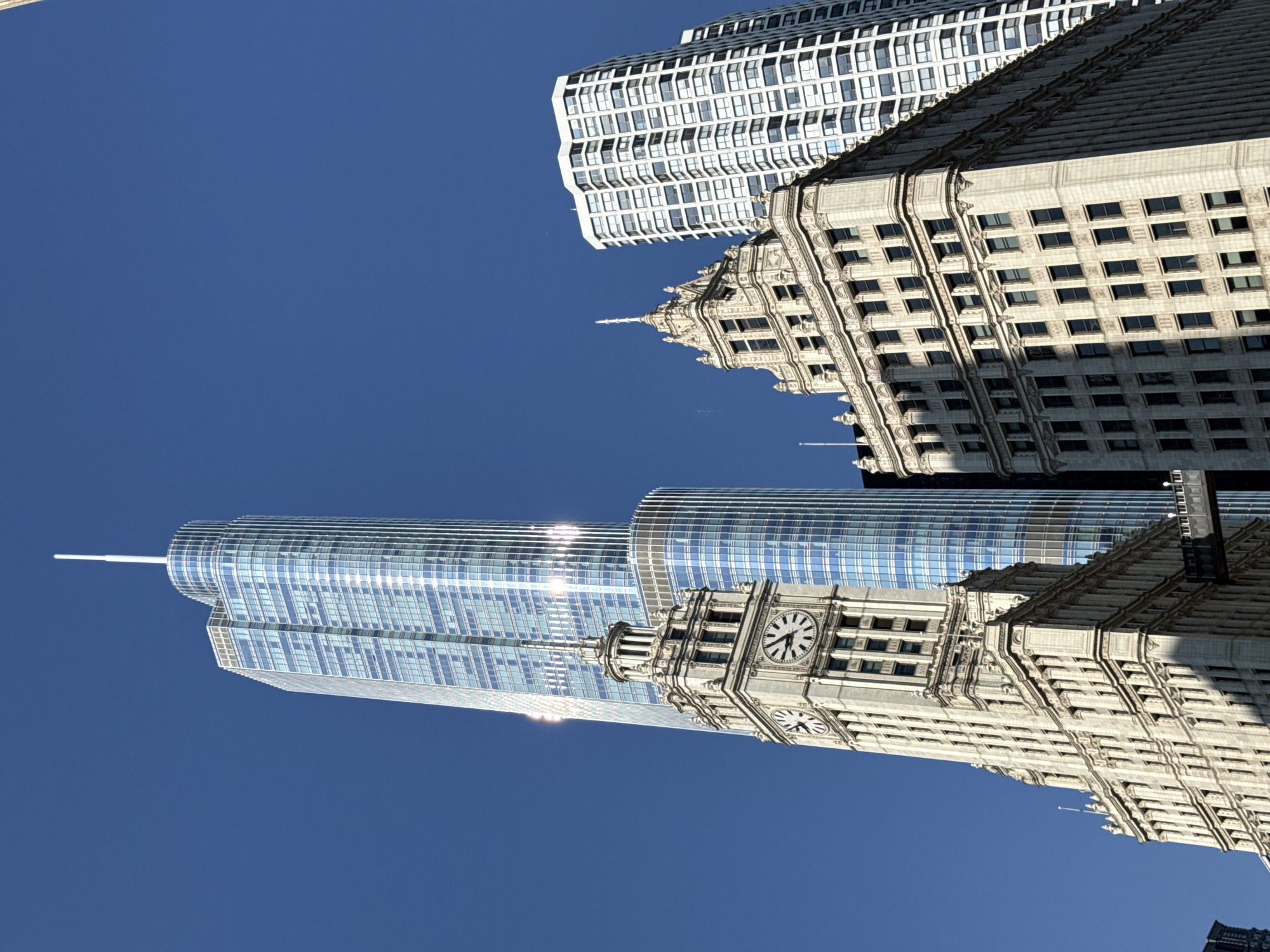 A low-angle, outdoor, eye-level shot showcases several distinct architectural styles of Chicago skyscrapers against a clear, bright blue sky. The tallest building on the left is a modern, slender skyscraper with a glass facade, reflecting the sunlight and appearing in shades of blue and white. To the left of this, a historic white stone tower with a clock face is prominently featured, its classical architectural details standing in contrast to the modern building. To the right of the modern skyscraper is another white stone building with ornate gothic-inspired architectural elements, including a pointed spire. Further to the right, a rectangular white building with a repeating pattern of windows is visible, representing a more contemporary but less elaborate design. A pedestrian bridge connects two buildings in the mid-ground. The image is devoid of any people, animals, or vehicles, focusing solely on the urban landscape and its impressive structures.