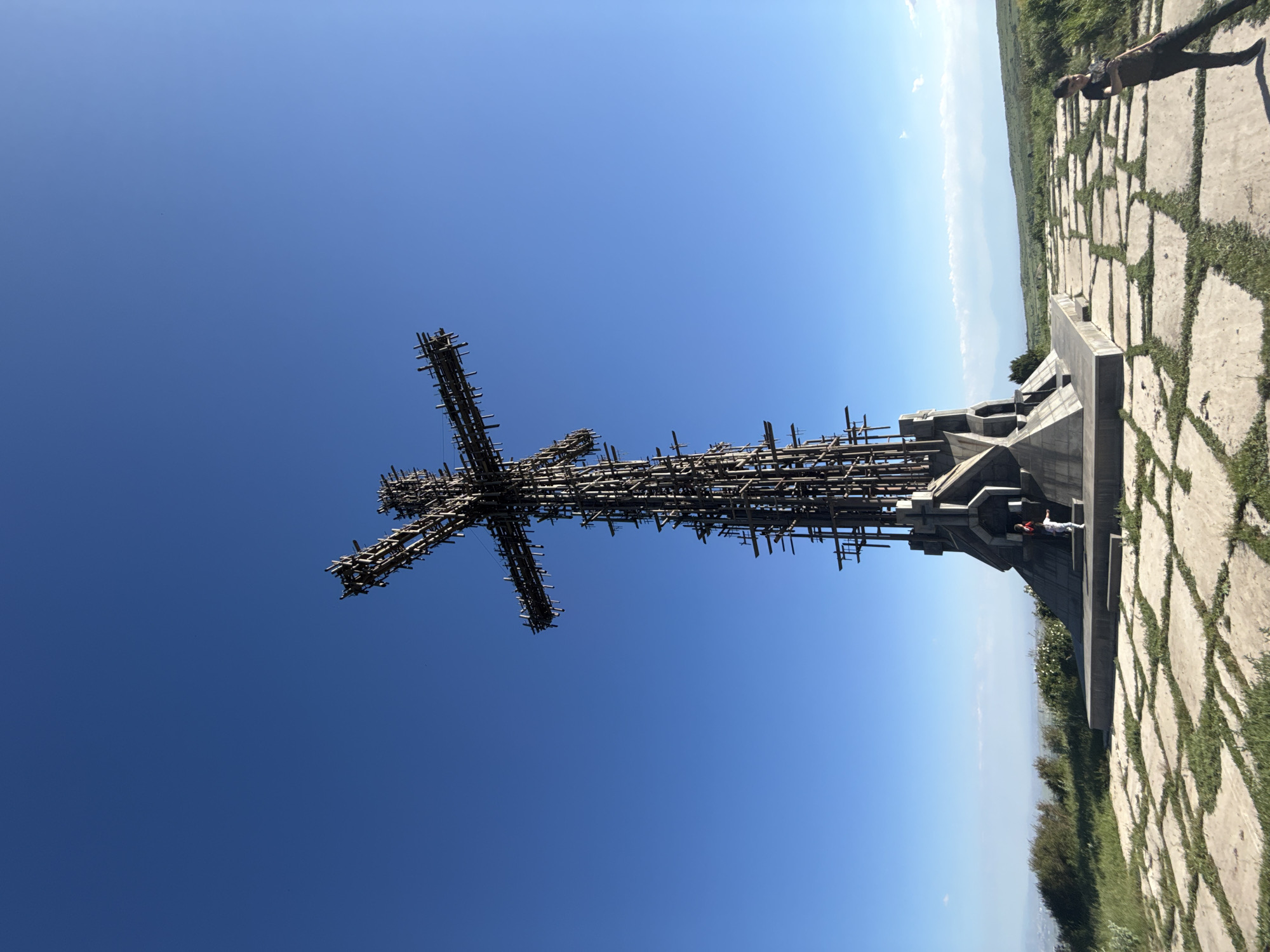 A large, industrial cross sculpture stands tall on a hilltop under a clear blue sky. The cross is constructed from a complex network of metal rods and beams, giving it a skeletal appearance. At the base of the cross is a concrete structure with angular, geometric shapes. Paved stone slabs with grass growing between them form a pathway leading to the sculpture. Two people are visible near the base of the cross, one a child in white and another adult in darker clothing. Further away, on the right side of the image, a young person in a dark t-shirt and pants leans against the pavement, looking towards the left. The surrounding landscape is hilly and covered with green grass and some vegetation. Distant mountains are visible on the horizon under a hazy sky.