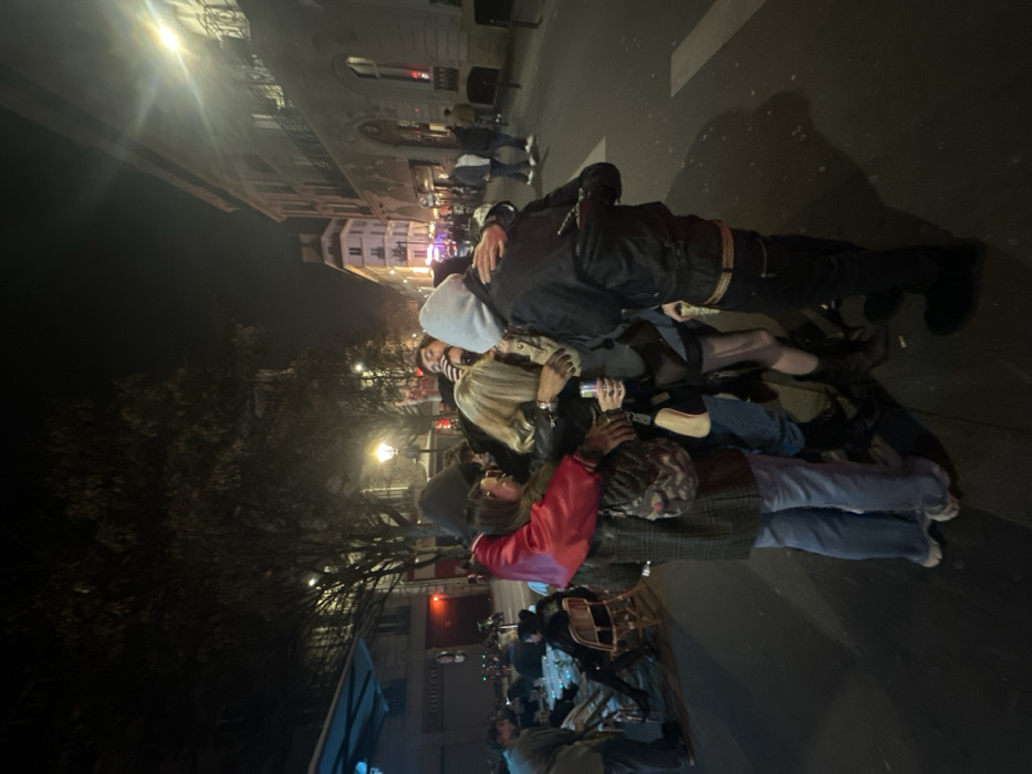 The image depicts a nighttime street scene in Paris, France, characterized by traditional Haussmannian architecture and an active urban environment. In the immediate foreground, a cluster of approximately five individuals stands in close physical interaction, appearing to be embracing or huddled together.
One person on the left wears a dark hoodie, a red-sleeved jacket, and light blue jeans, with their arm around another individual. A central figure with blonde hair is seen holding a silver beverage can. To the right, another person wears a light grey hoodie, a black jacket, and dark trousers, with their arm extended towards the central blonde figure. Partially visible individuals are intertwined within this group, including one making a "rock on" hand gesture in the background.
To the left of the main group, an outdoor seating area of a commercial establishment, possibly a café or restaurant, is visible under a blue-lit awning. Several patrons are seated at small round tables with chairs. The street, partially illuminated by artificial light sources including streetlights, recedes into the background where additional pedestrians are observed walking. Buildings with distinct Parisian architectural features, such as multiple stories and ornate balconies, line the right side of the street. The ground is asphalt with a visible white pedestrian crossing stripe.